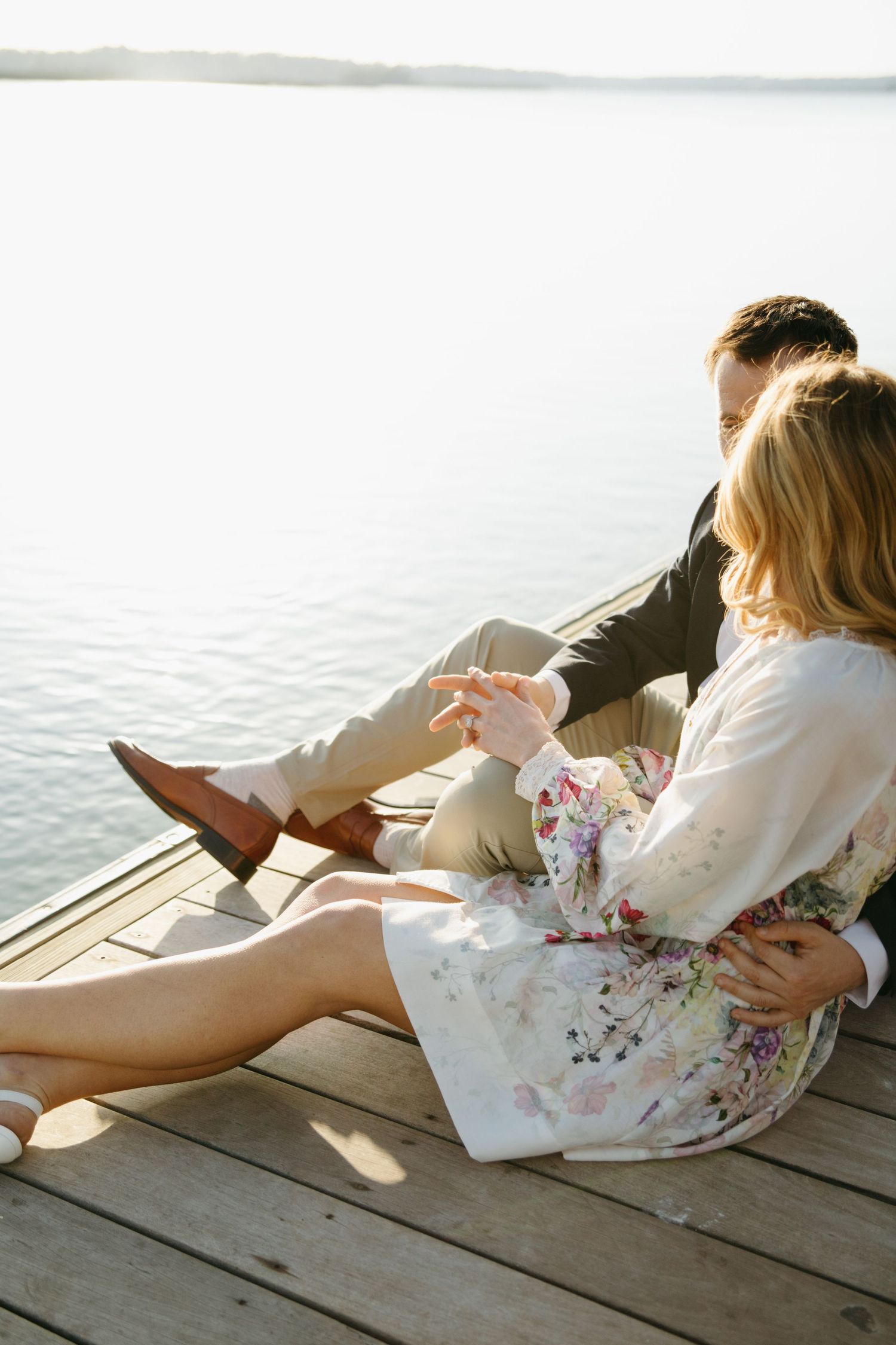 Couple sitting together on a dock overlooking a calm sunlit lake in casual attire.