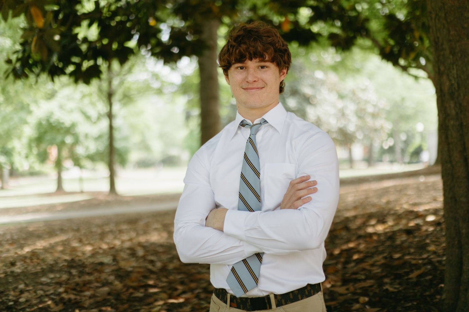 Young man with curly hair in white shirt and striped tie stands with arms crossed under trees.