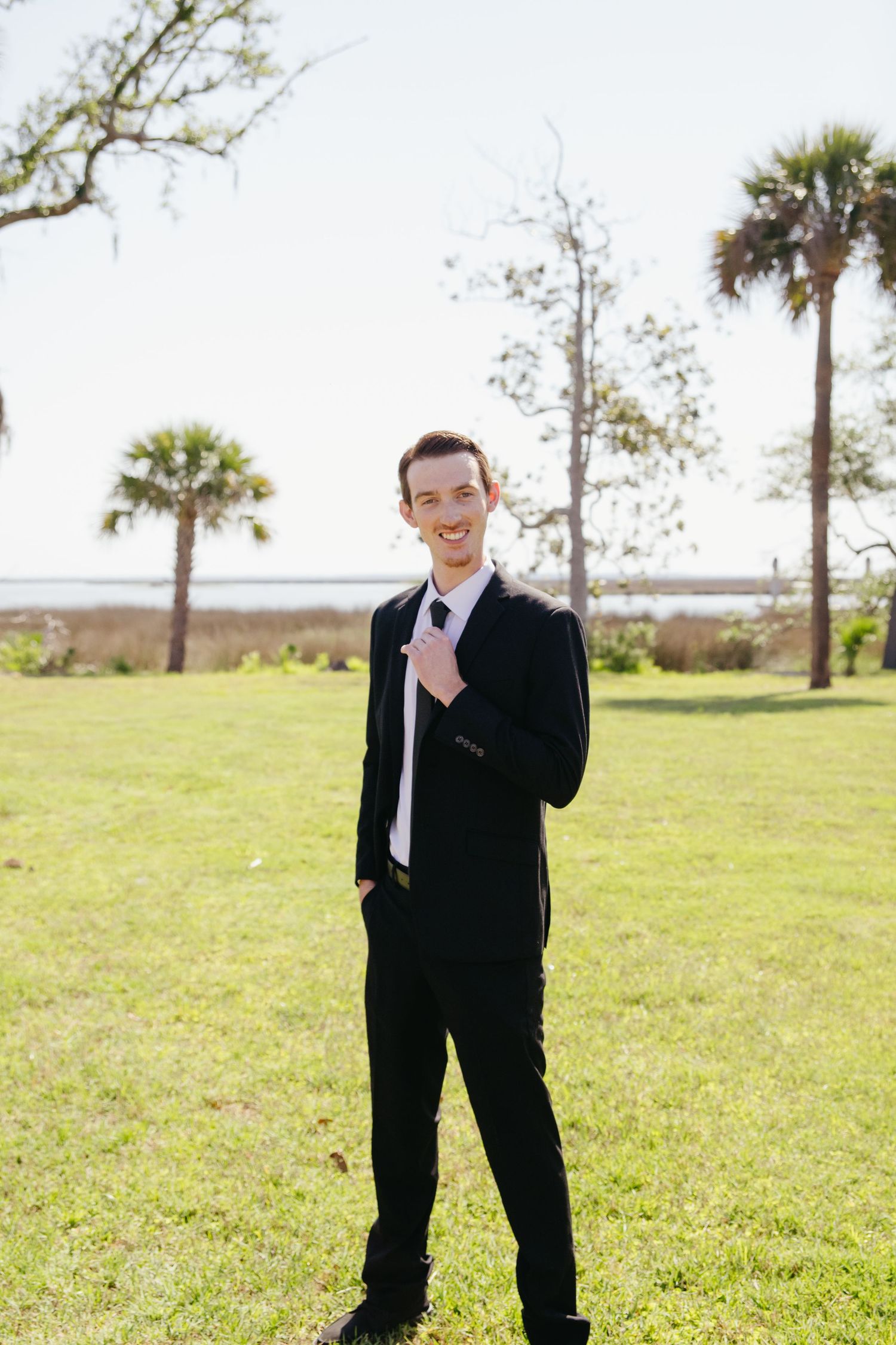 Smiling young man in black suit and white tie posing on green lawn with palm trees behind him.