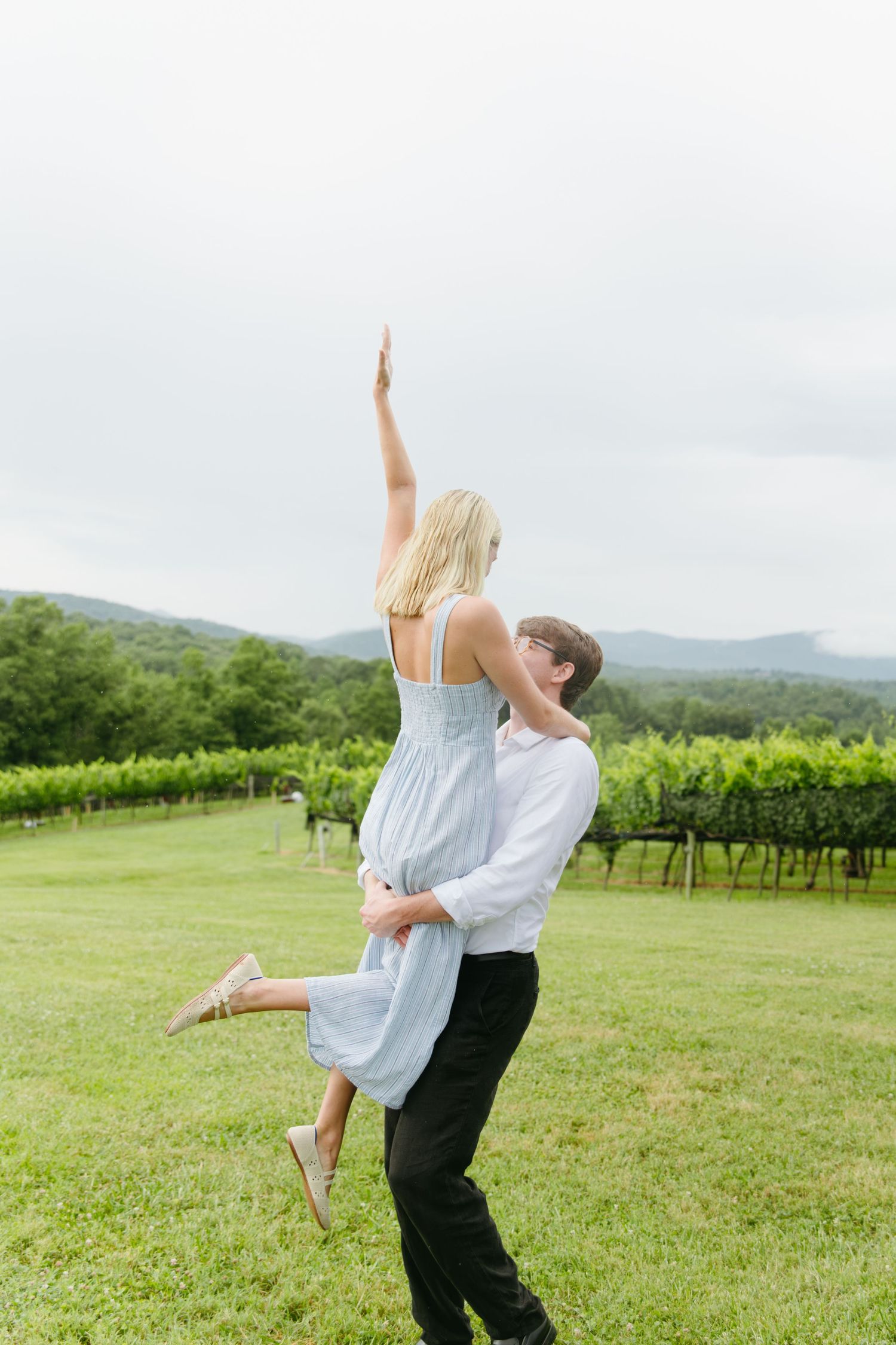 Man lifting woman in blue dress at vineyard engagement shoot with mountain views.