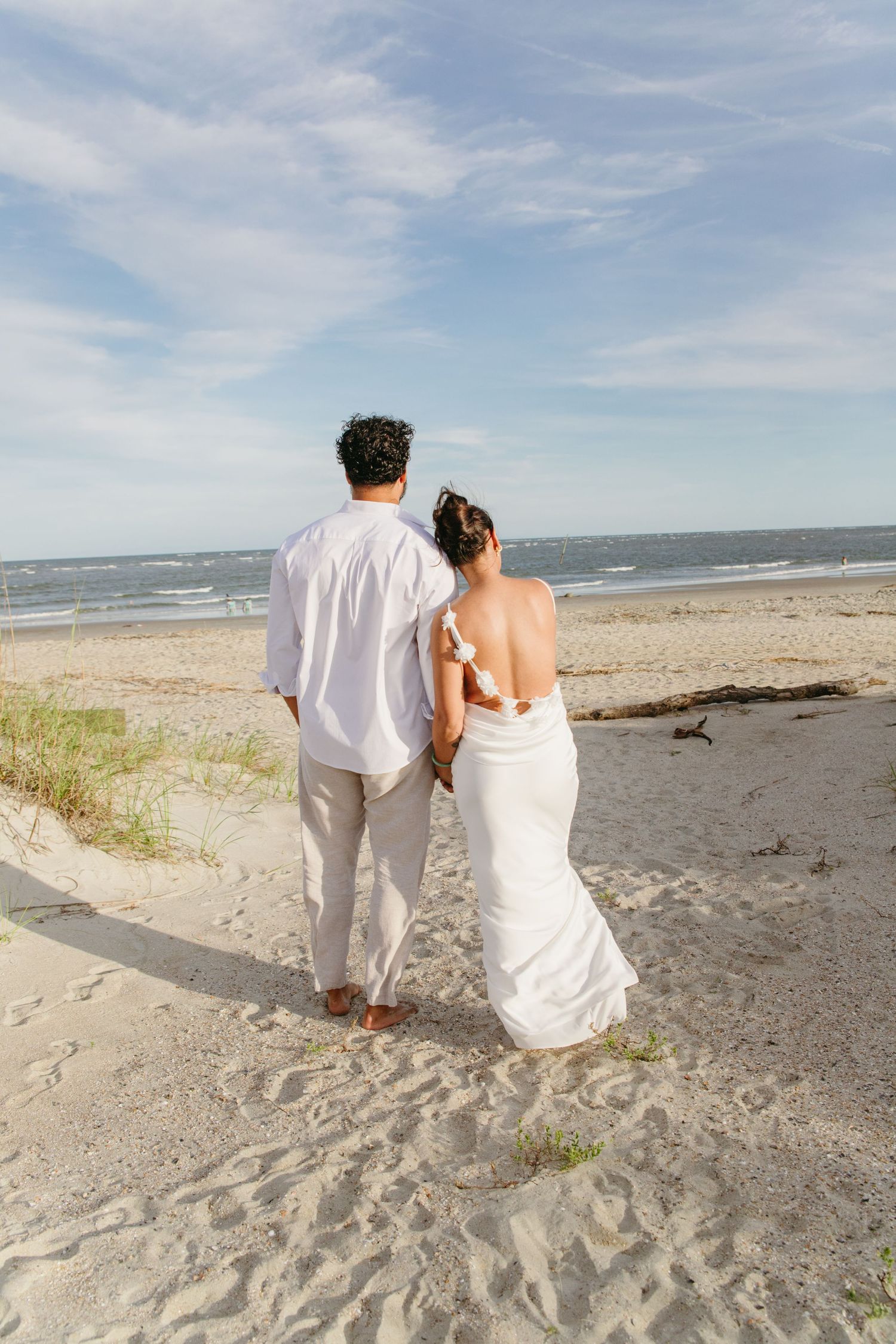 Couple in white holding hands walking toward ocean on sandy beach from behind.