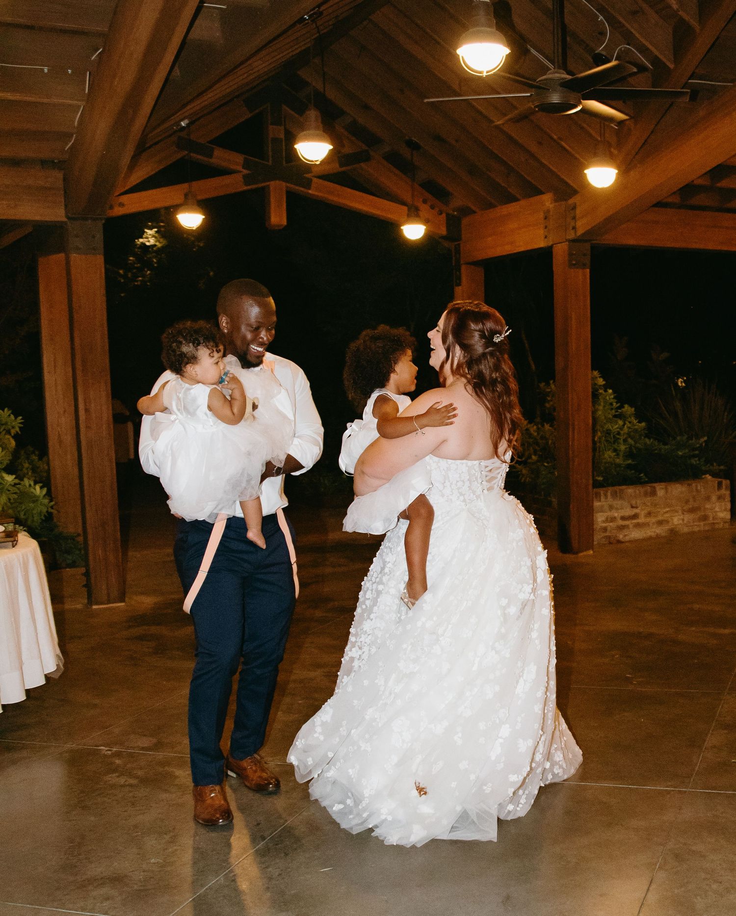 Bride and groom each holding a child while dancing together under pavilion lights at wedding reception.