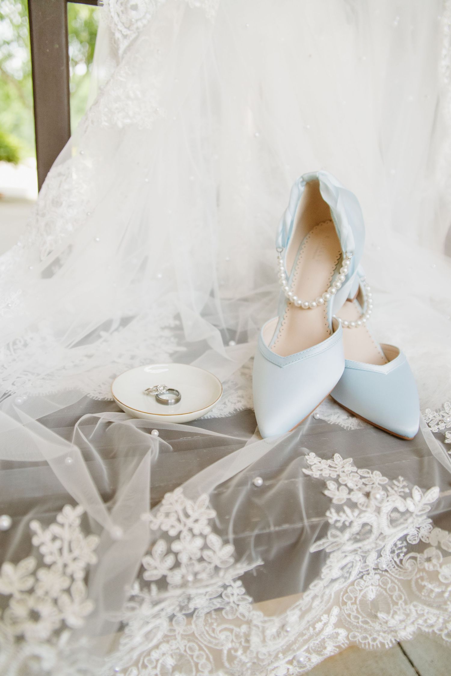 Light blue bridal heels with pearl anklet strap beside wedding rings on lace veil.