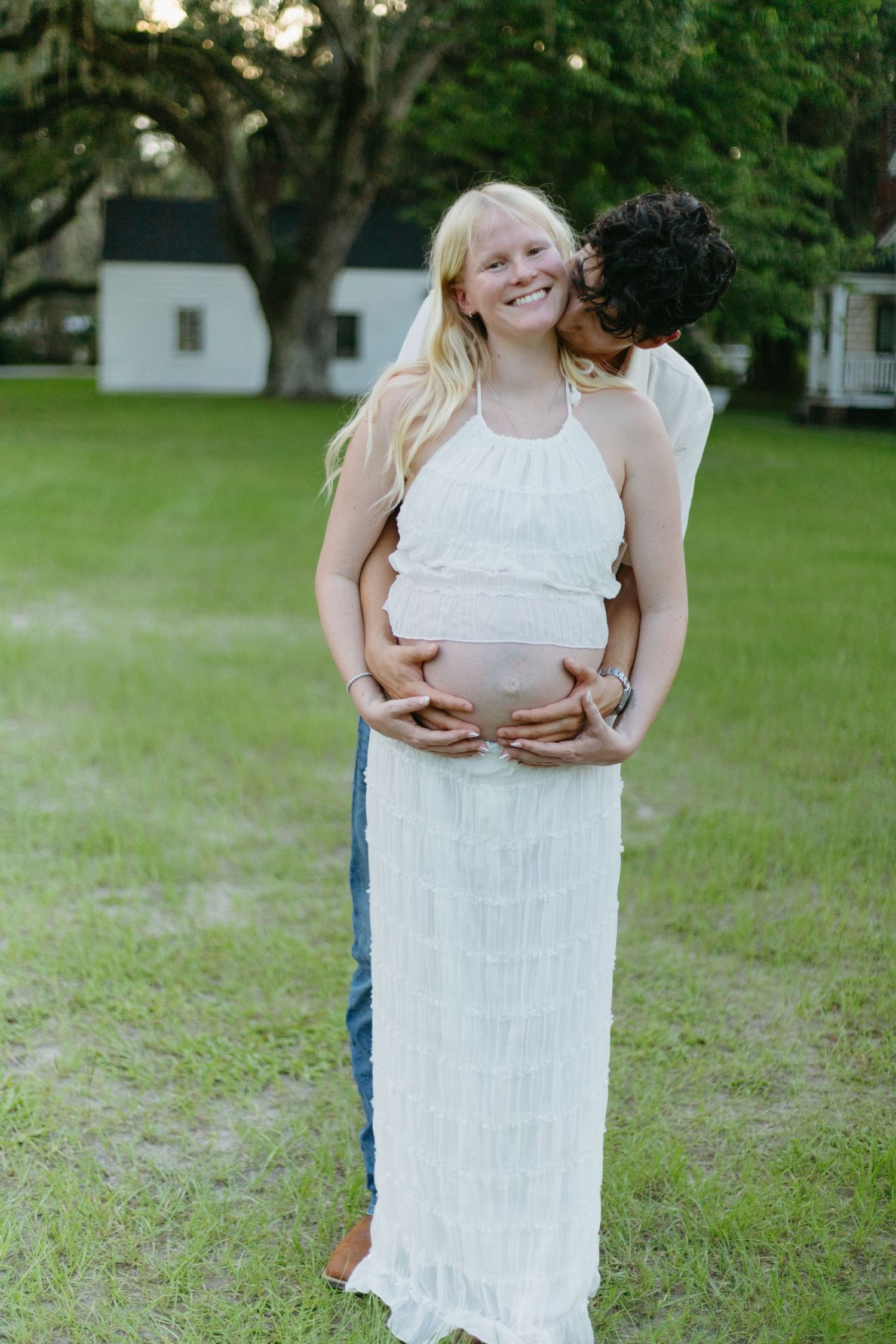 Glowing pregnant woman in white dress being embraced from behind by partner outdoors.