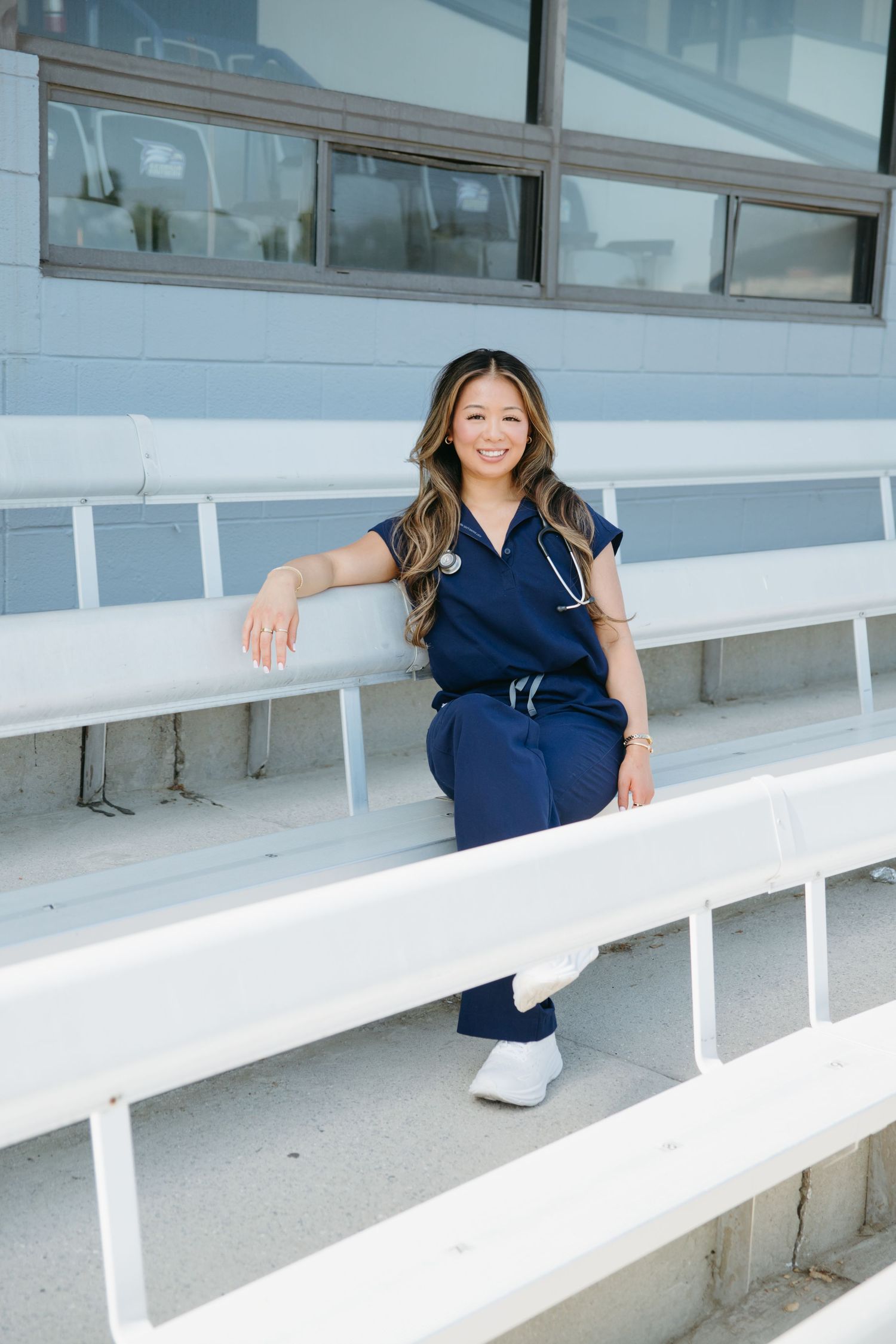 Asian woman in navy blue scrubs with stethoscope sitting on bleachers smiling.