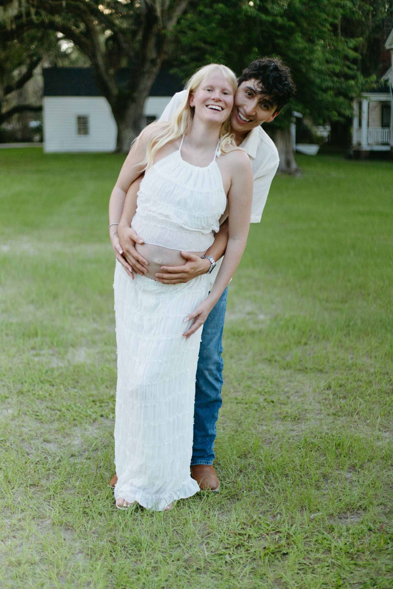 Smiling pregnant woman in white dress embraced by partner in a green outdoor setting.