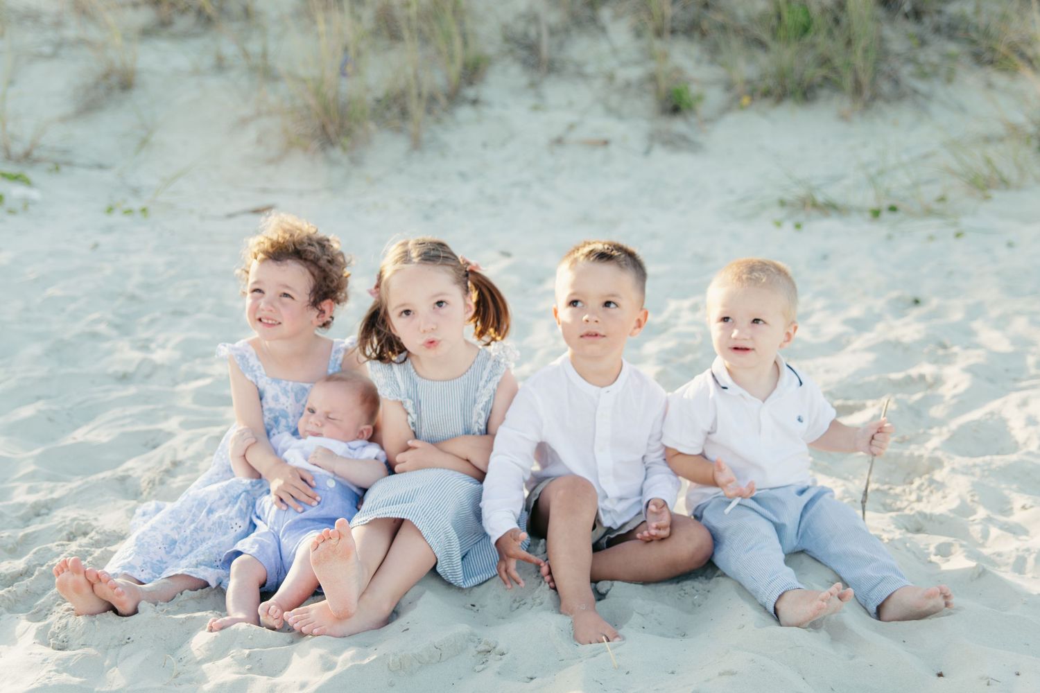 Five young children sit together on sandy beach dunes, dressed in light blue and white outfits, with the youngest as a newborn.