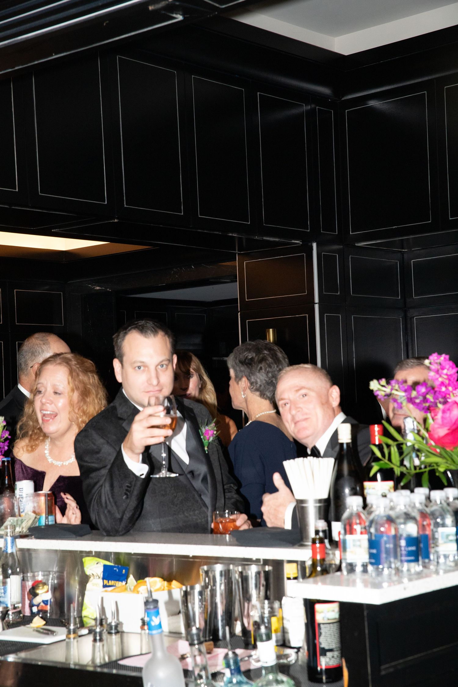 Wedding guests celebrating at sleek black bar, groom raising drink in formal attire.