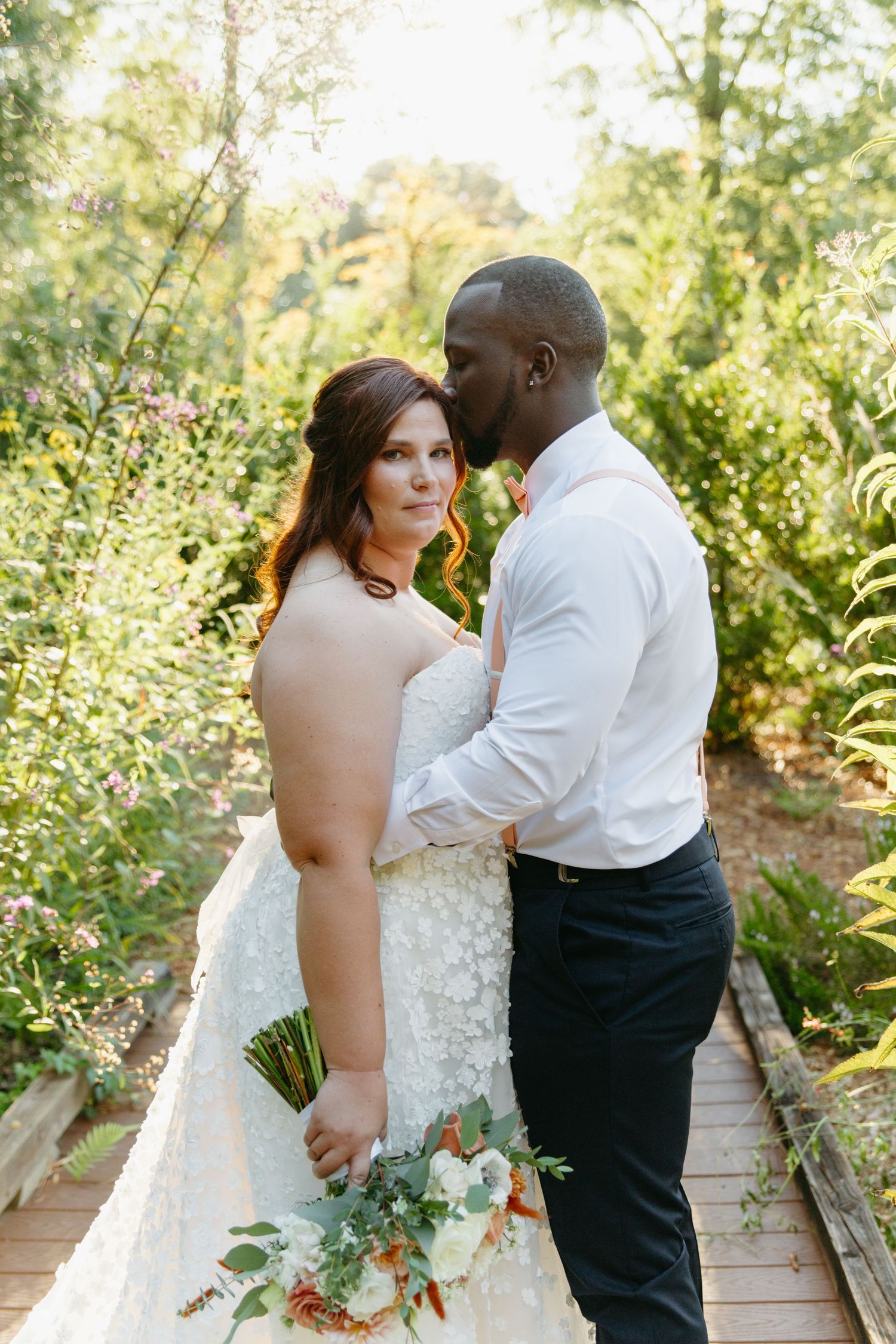 Interracial couple sharing a tender moment on a wooden bridge surrounded by lush garden greenery.