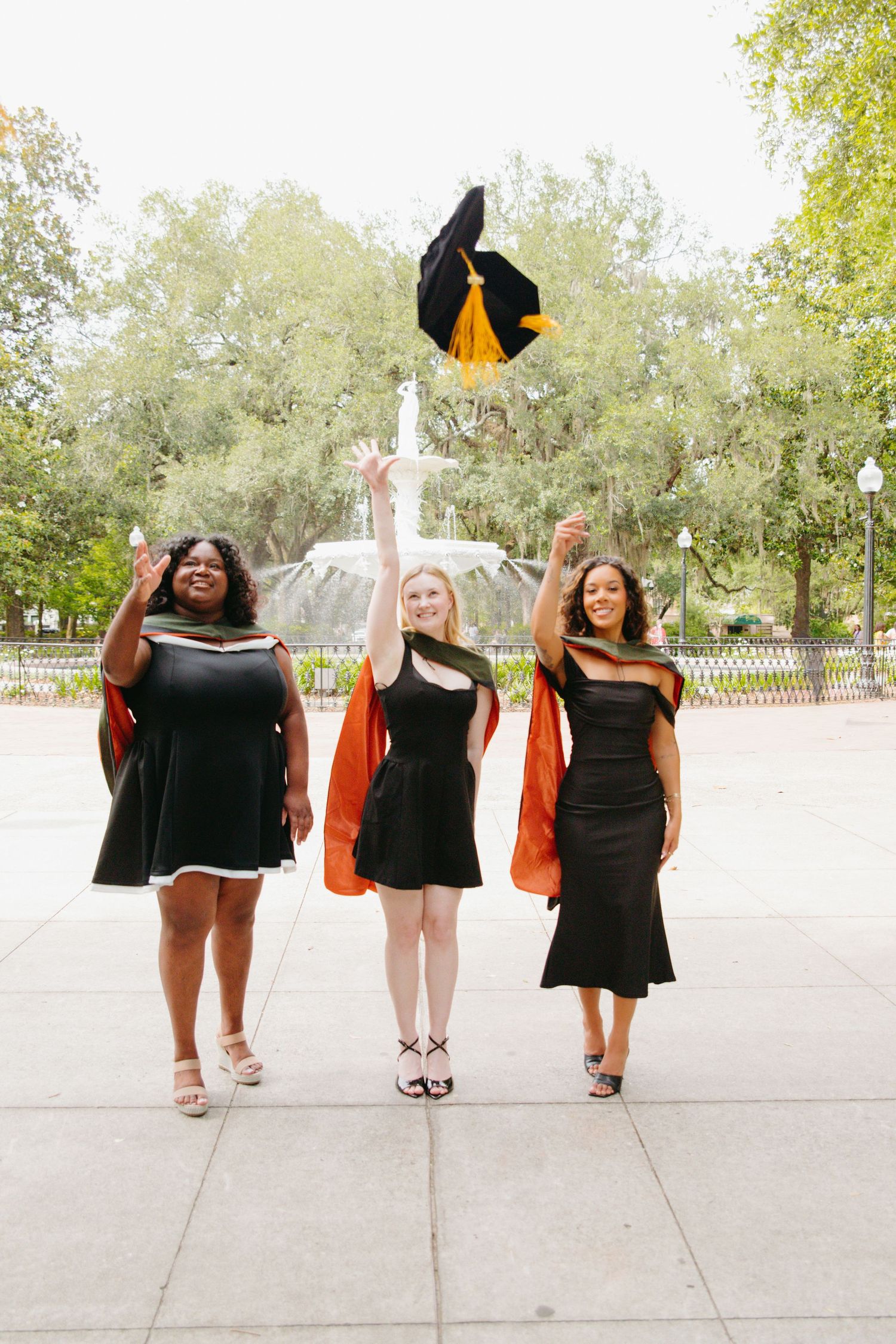 Three female graduates in black dresses celebrate by tossing caps near fountain.