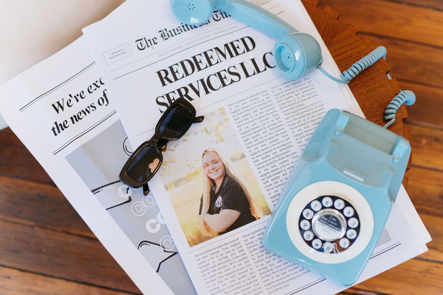 Redeemed Services LLC newspaper feature with vintage blue rotary phone, sunglasses, and business photo on wooden surface.