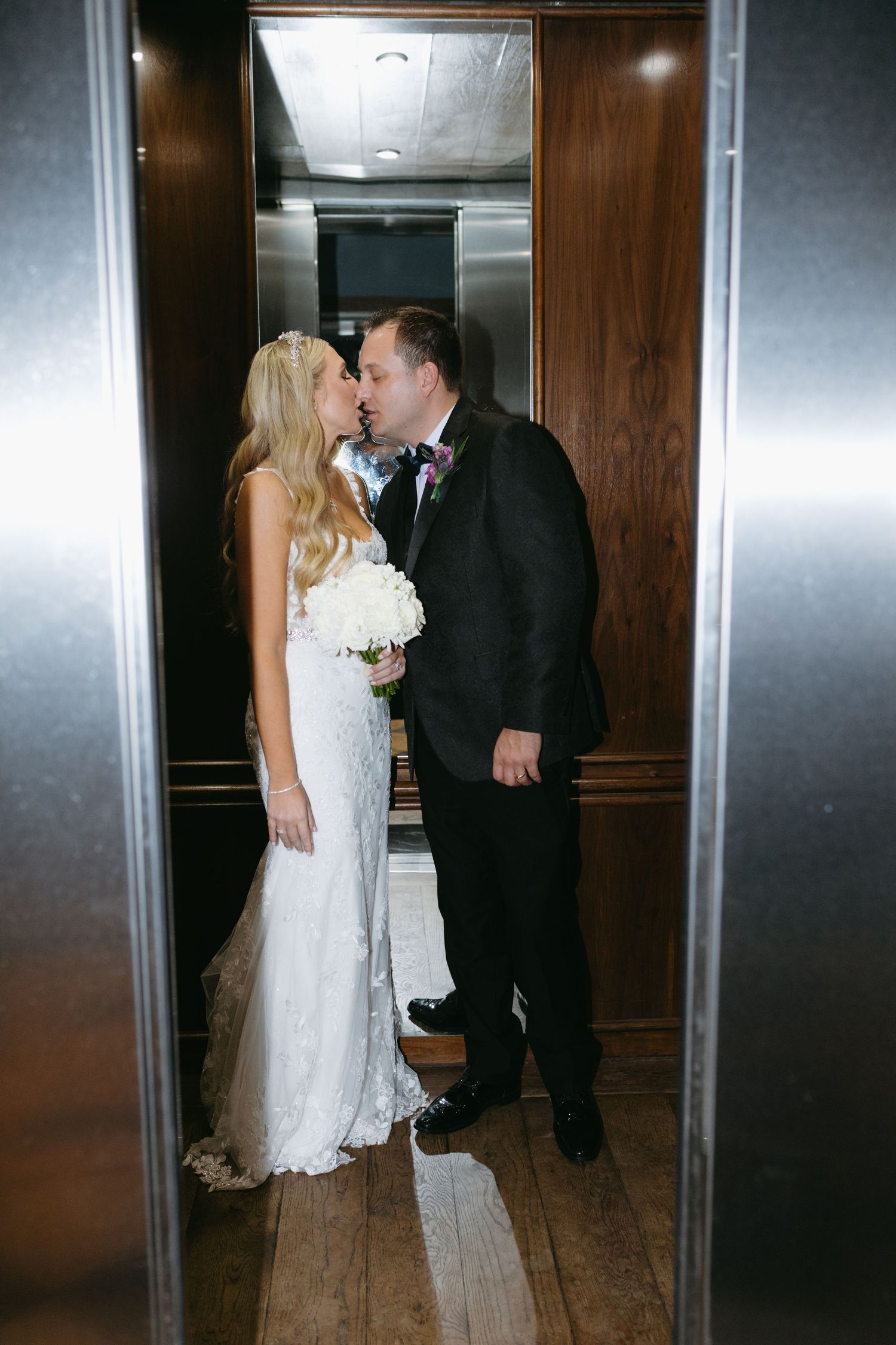 Bride and groom sharing a kiss inside an elevator, bride holding white floral bouquet.