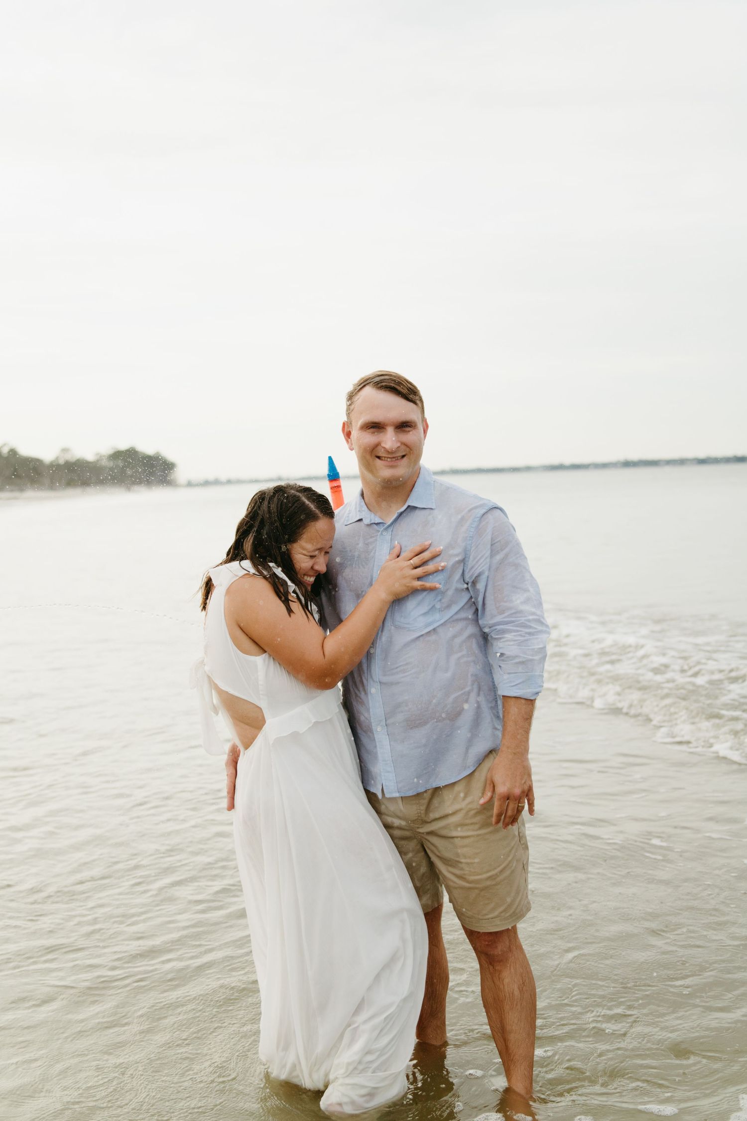 Couple stands embracing in ocean water, smiling, on an overcast beach day.