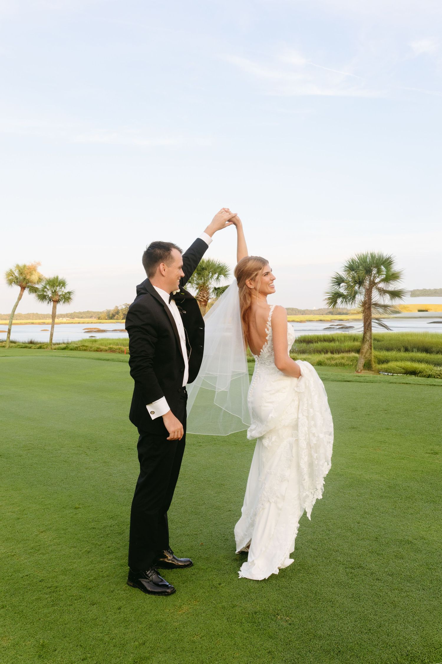 Groom twirls bride in backless lace gown on golf course with palm trees and marsh views at sunset.