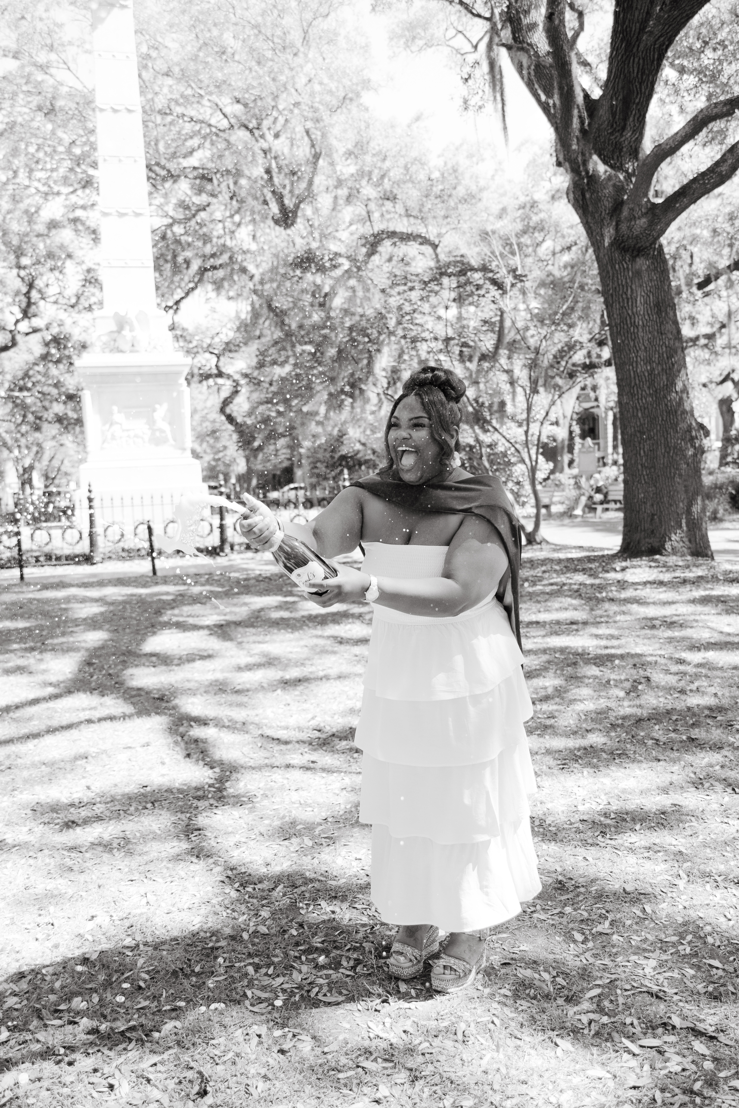 Black and white photo of joyful woman in white dress celebrating outdoors in park.