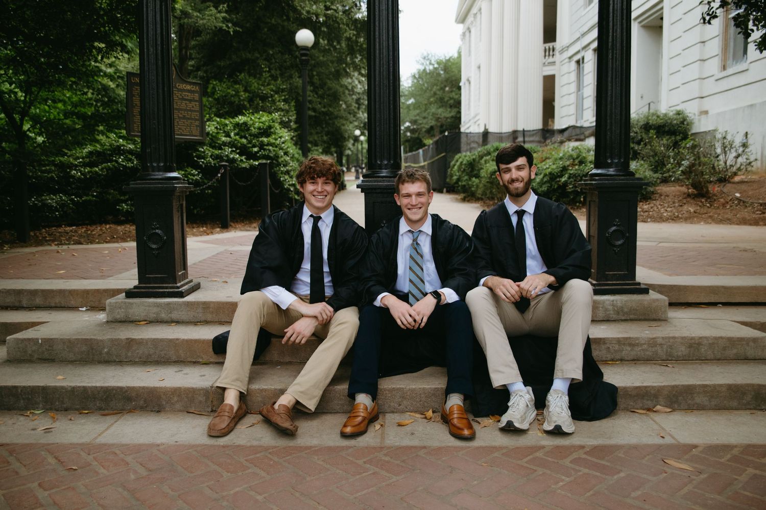 Three young men in blazers and ties sitting on brick steps near campus landmark for graduation photos.
