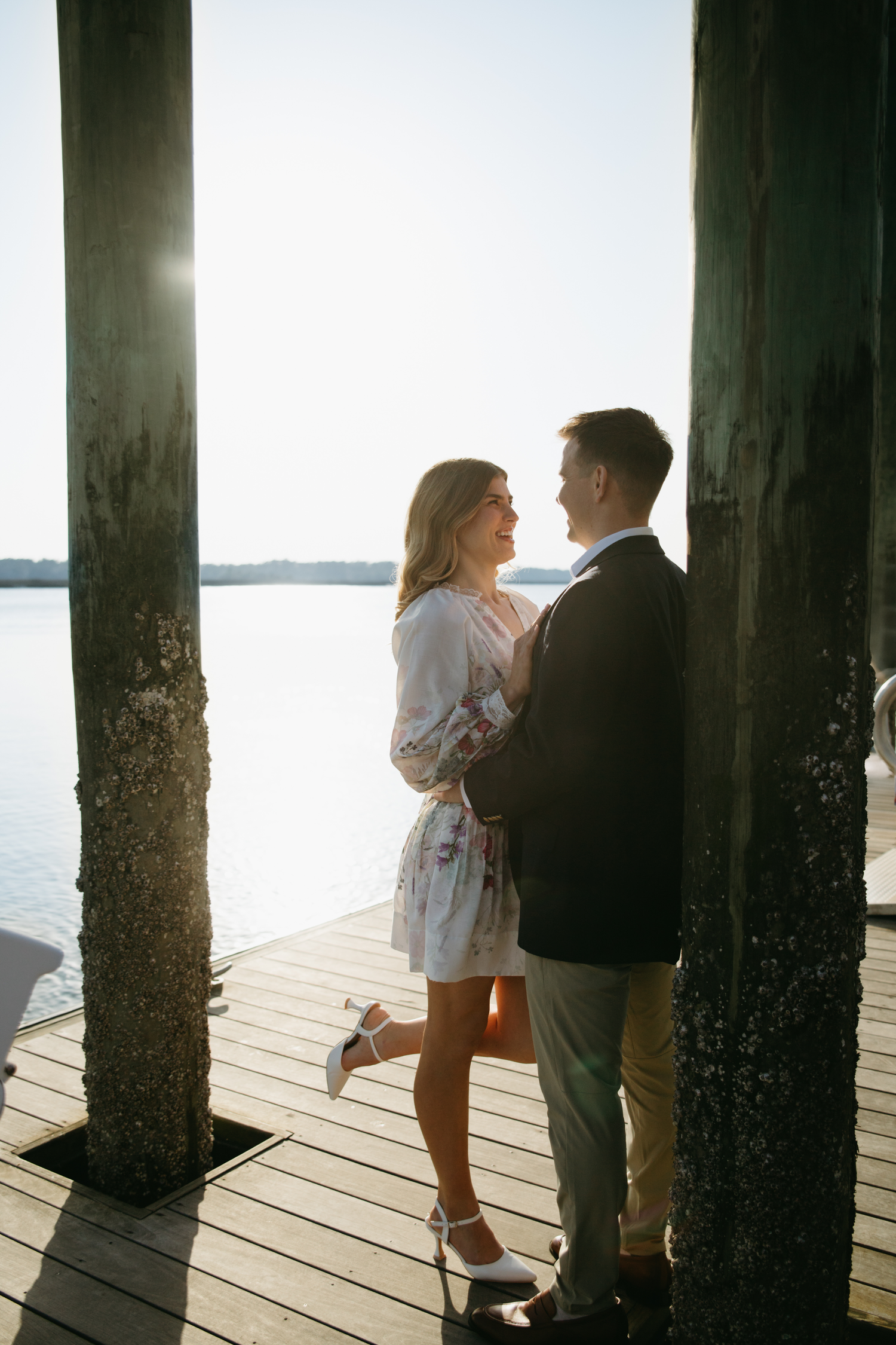 Couple embracing on a sunlit wooden dock by the water, woman in floral dress lifting her leg romantically.