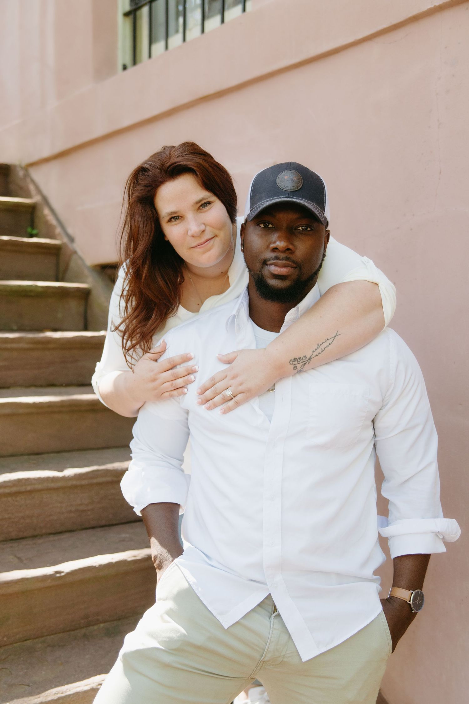 Interracial couple posing on outdoor steps, woman embracing man from behind against a pink wall.