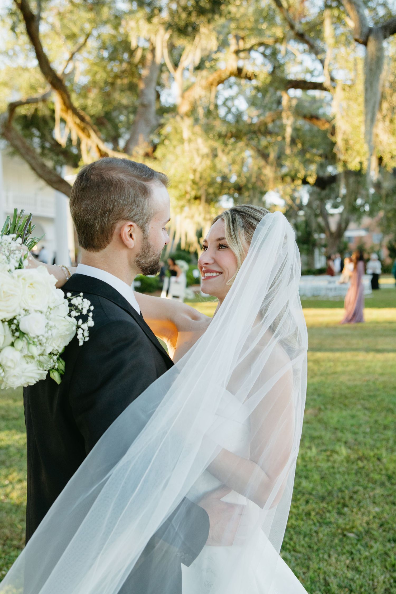 Radiant bride smiles at groom while holding white bouquet beneath sprawling oak trees at golden hour.