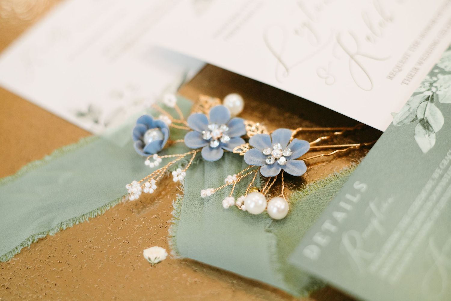 Blue floral hair pins with pearls on gold wedding stationery with sage green ribbon.