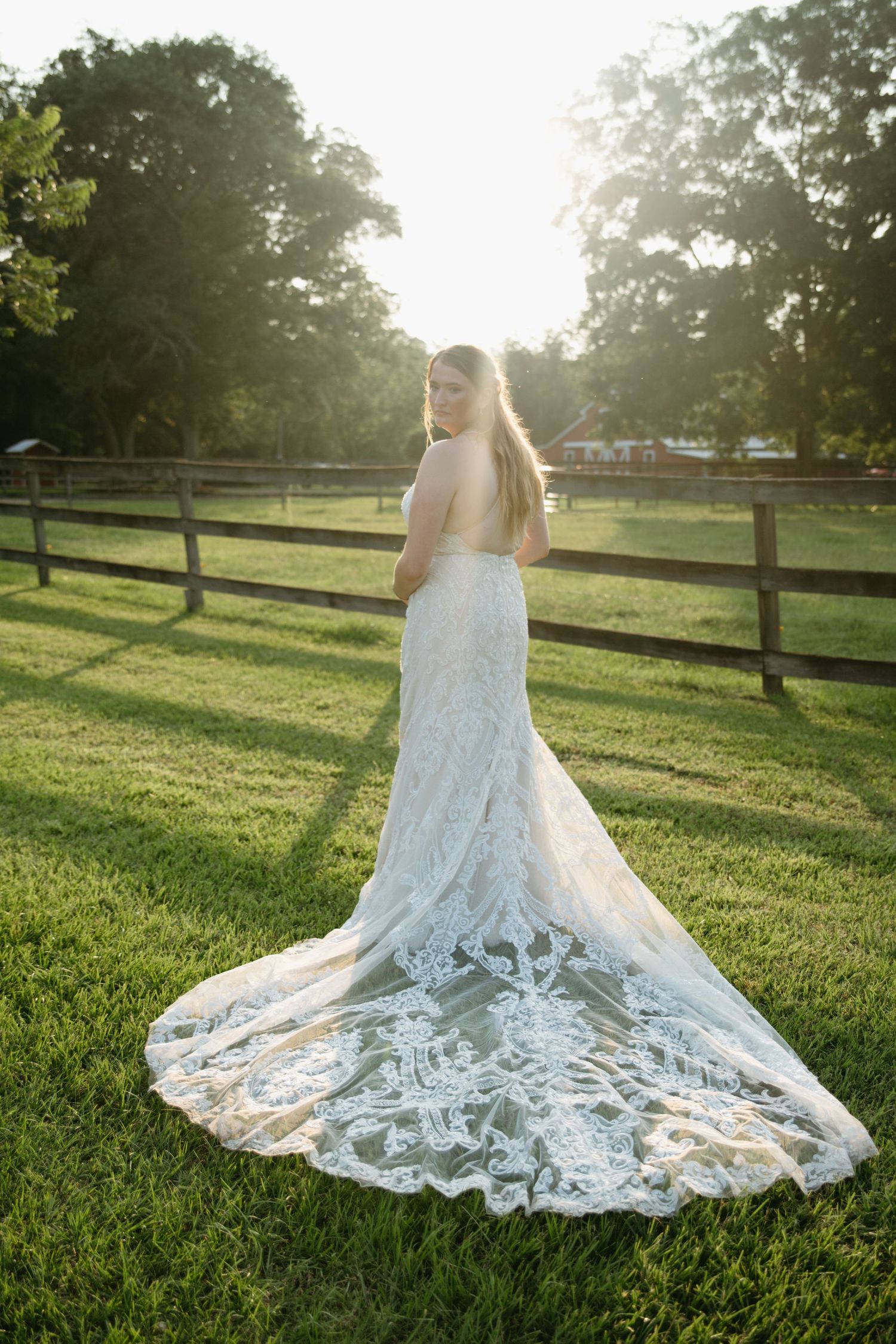 Bride in stunning lace wedding gown with long train poses in golden sunset light by fence.