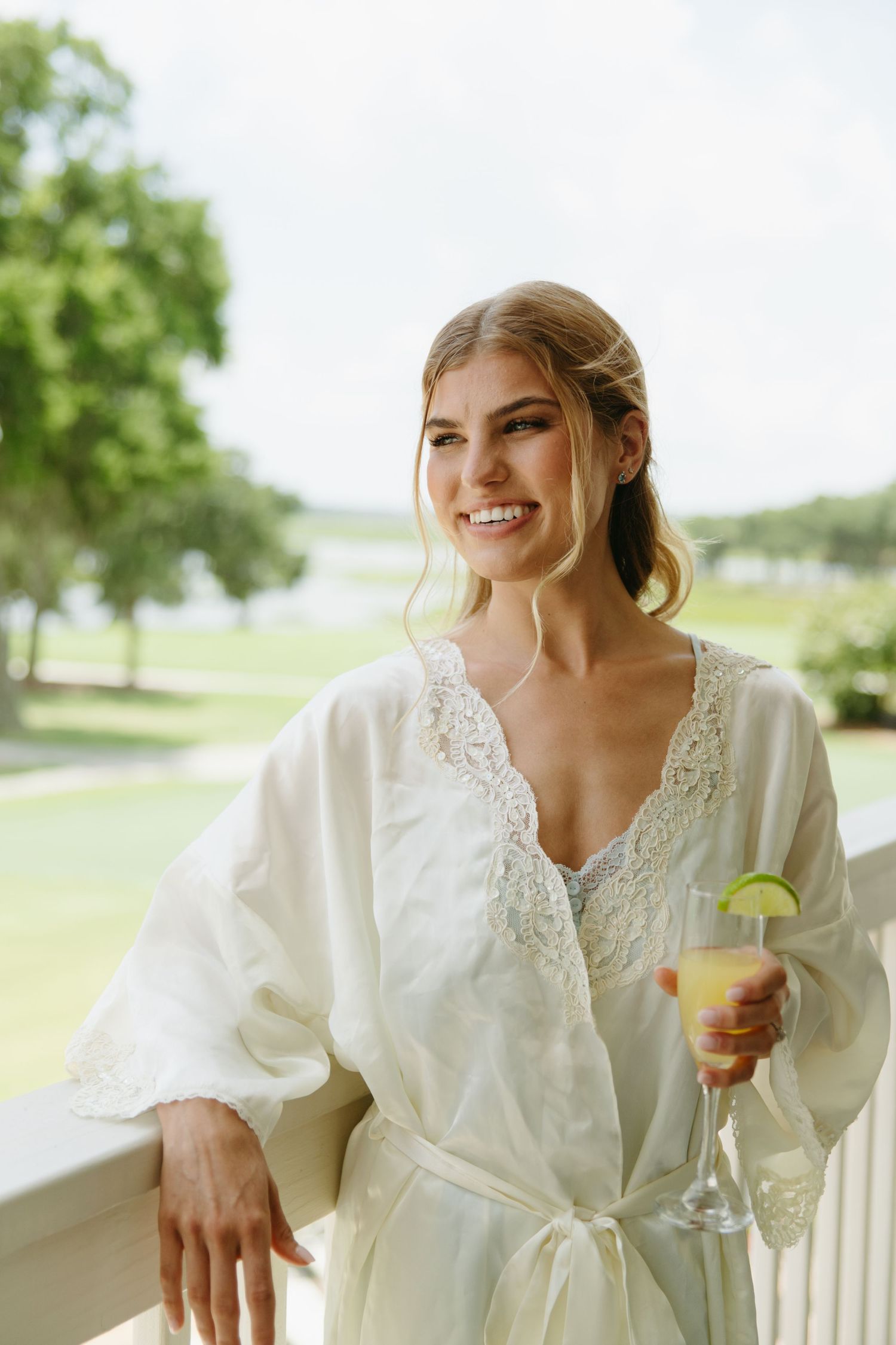 Smiling bride in white lace robe holds cocktail on outdoor balcony with green landscape behind her.