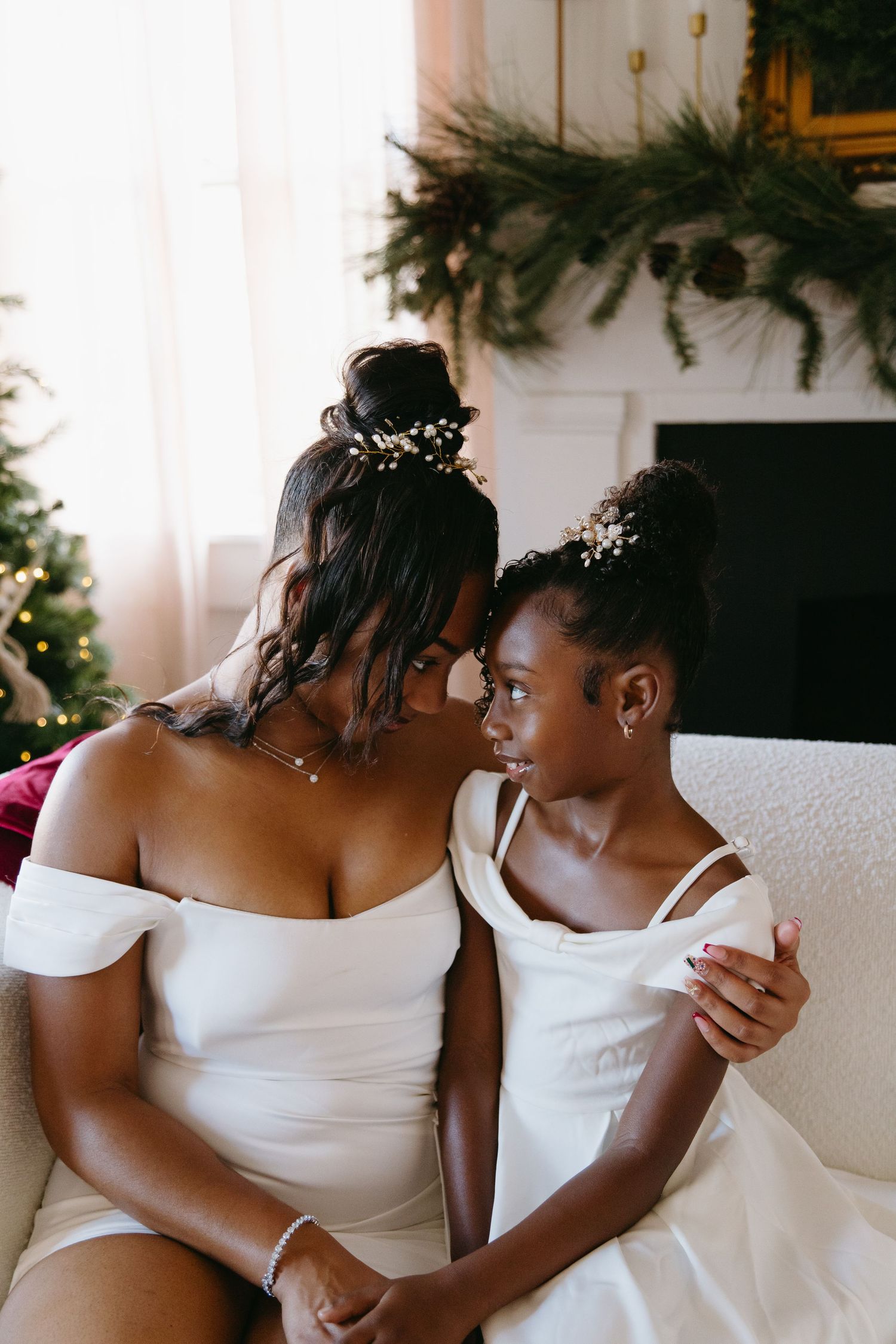 A mother and daughter in matching white dresses touch foreheads tenderly, both wearing floral hair accessories near a Christmas tree.