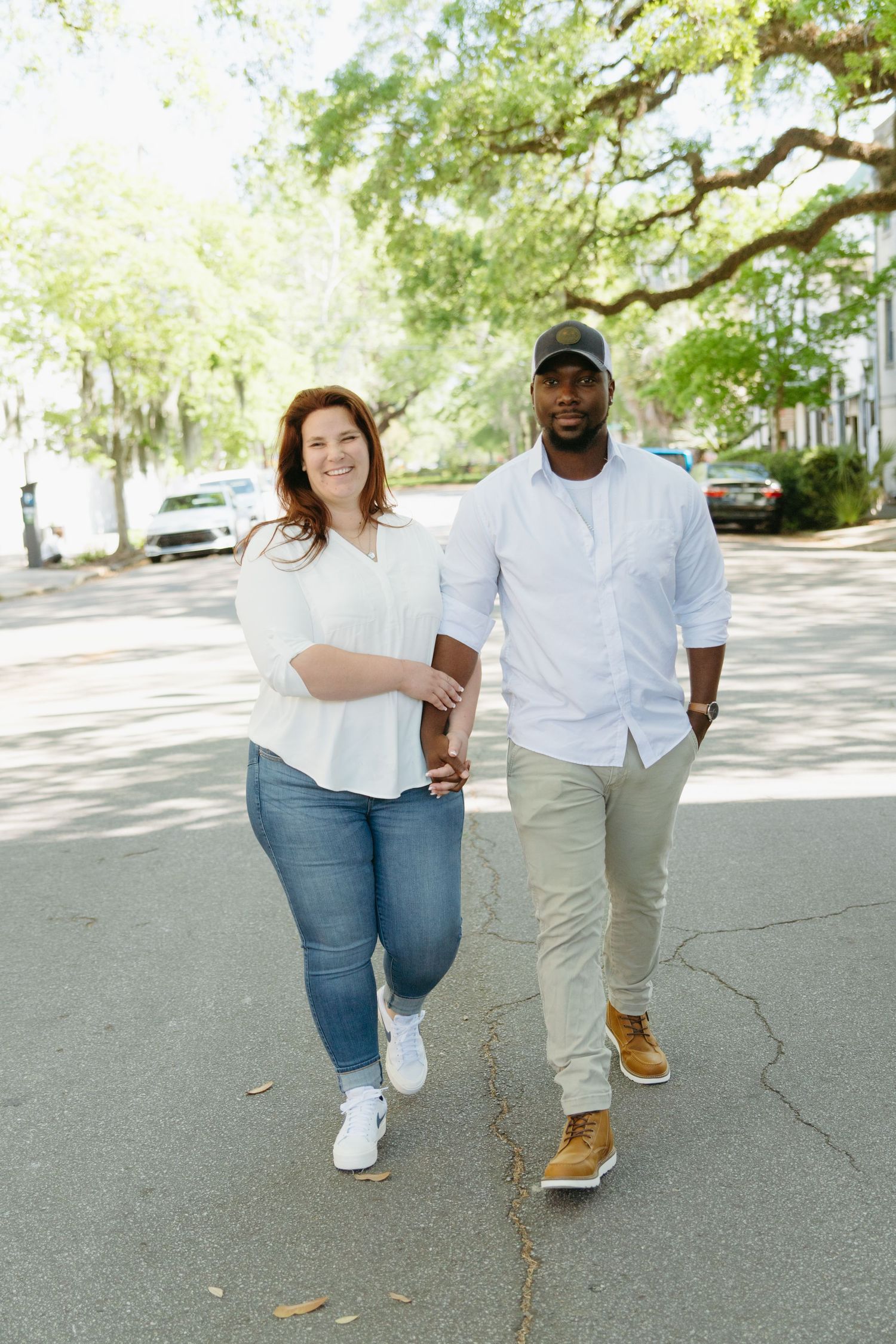 Interracial couple walking hand in hand down a tree-lined street on a sunny day.