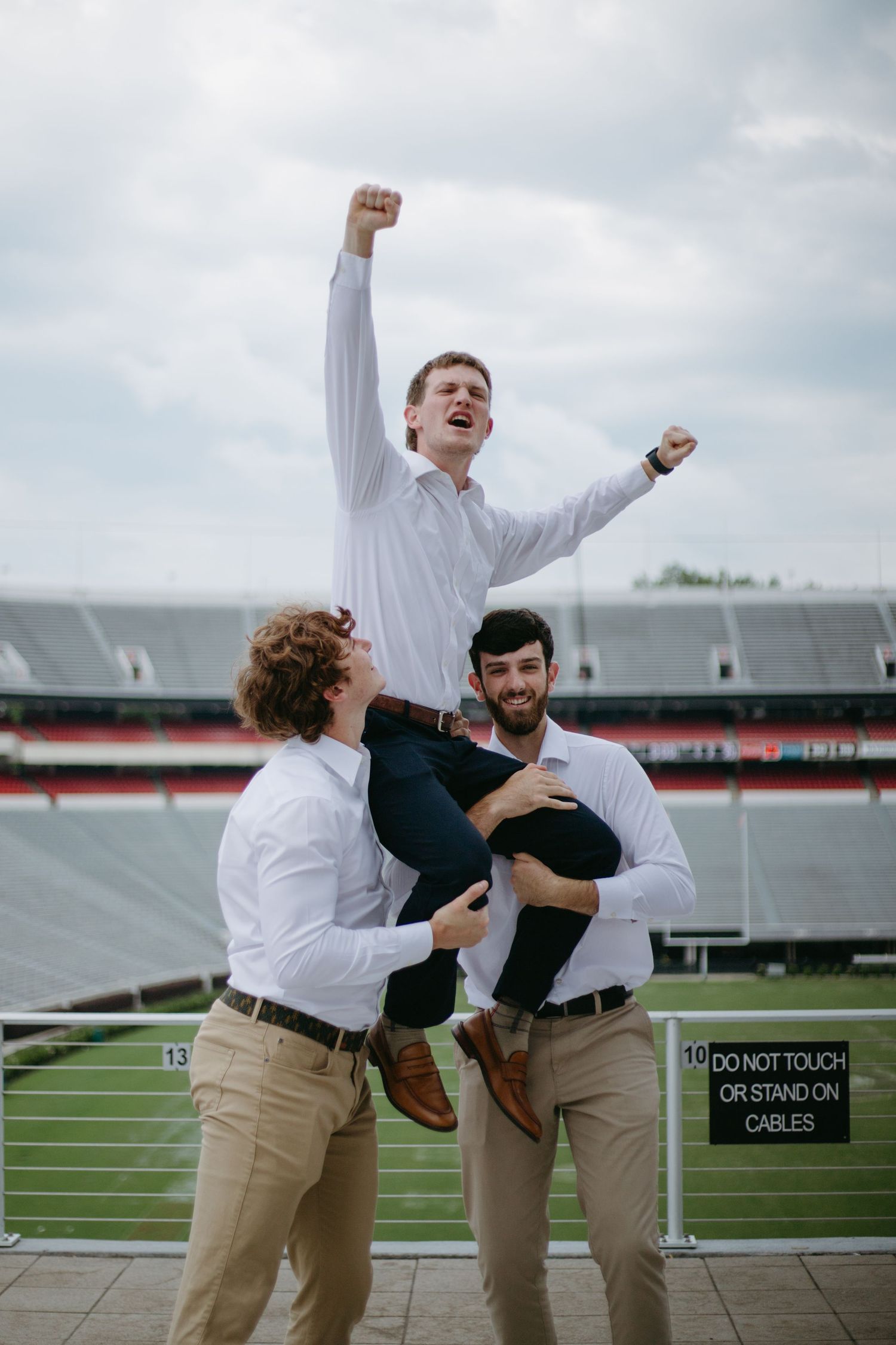 Three male graduates celebrating at stadium, two lifting third friend triumphantly in the air.