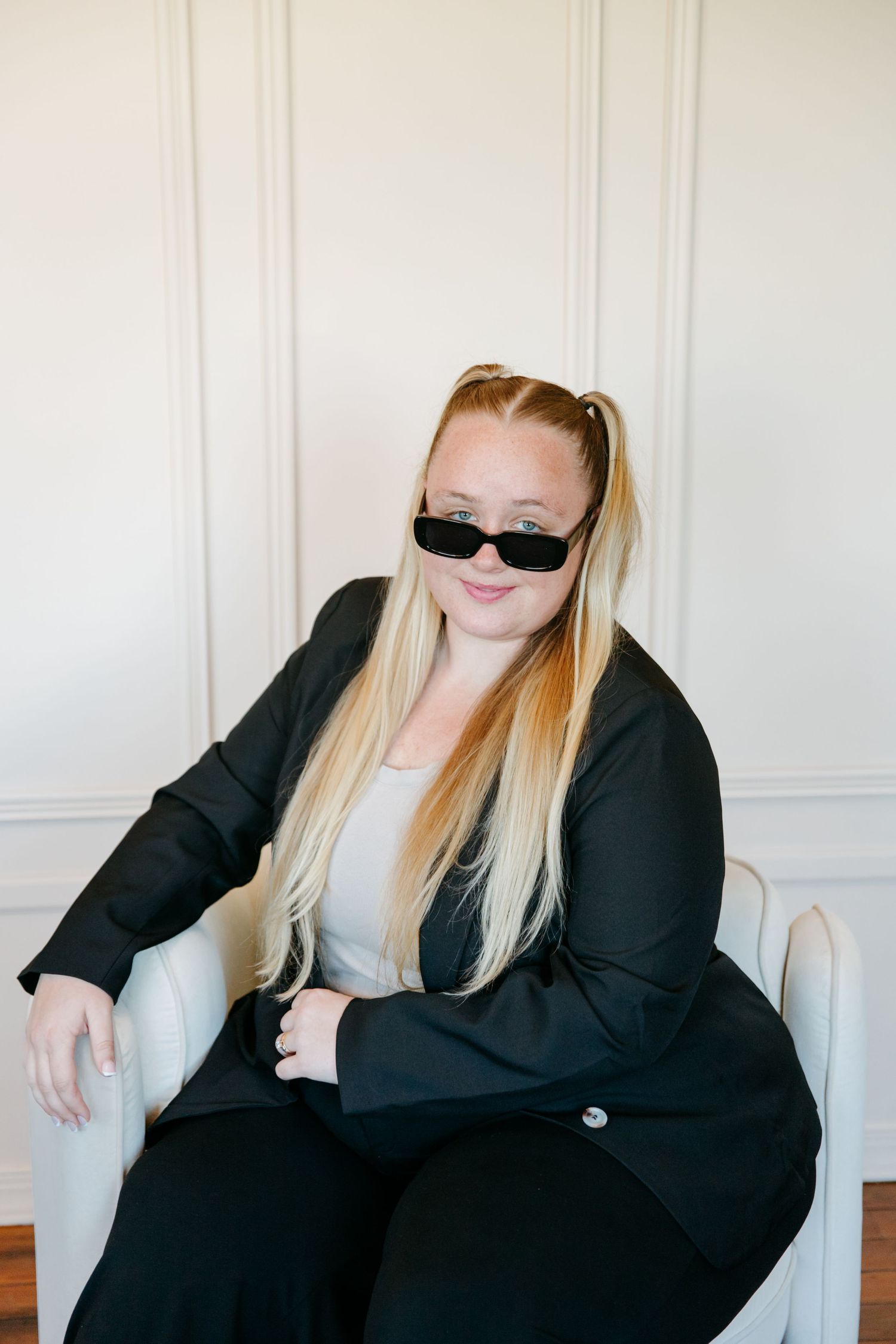 Confident blonde woman in black blazer and sunglasses sitting in white chair for professional business portrait.