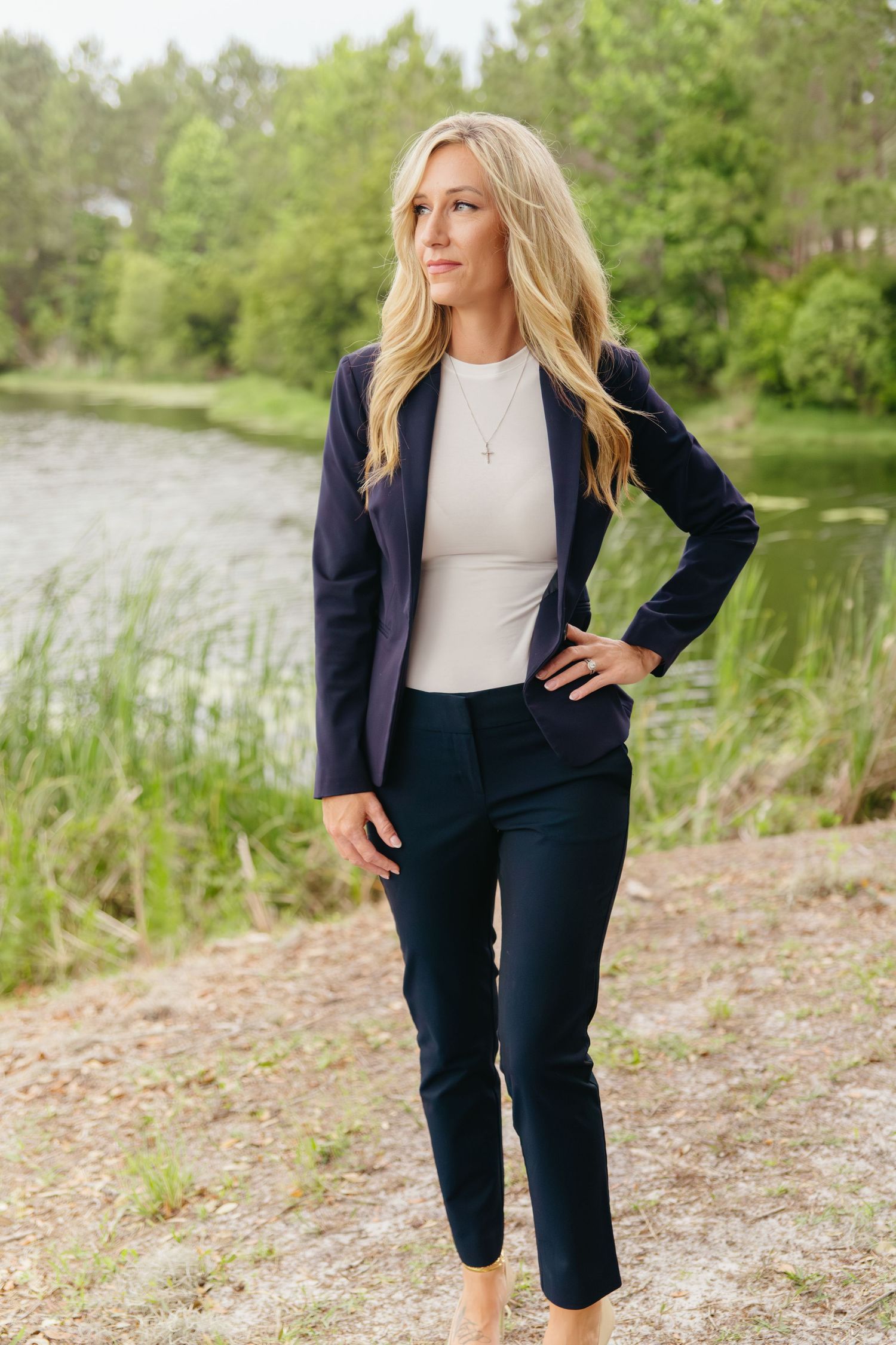 Professional blonde woman in navy blue blazer and pants posing outdoors near a serene pond.