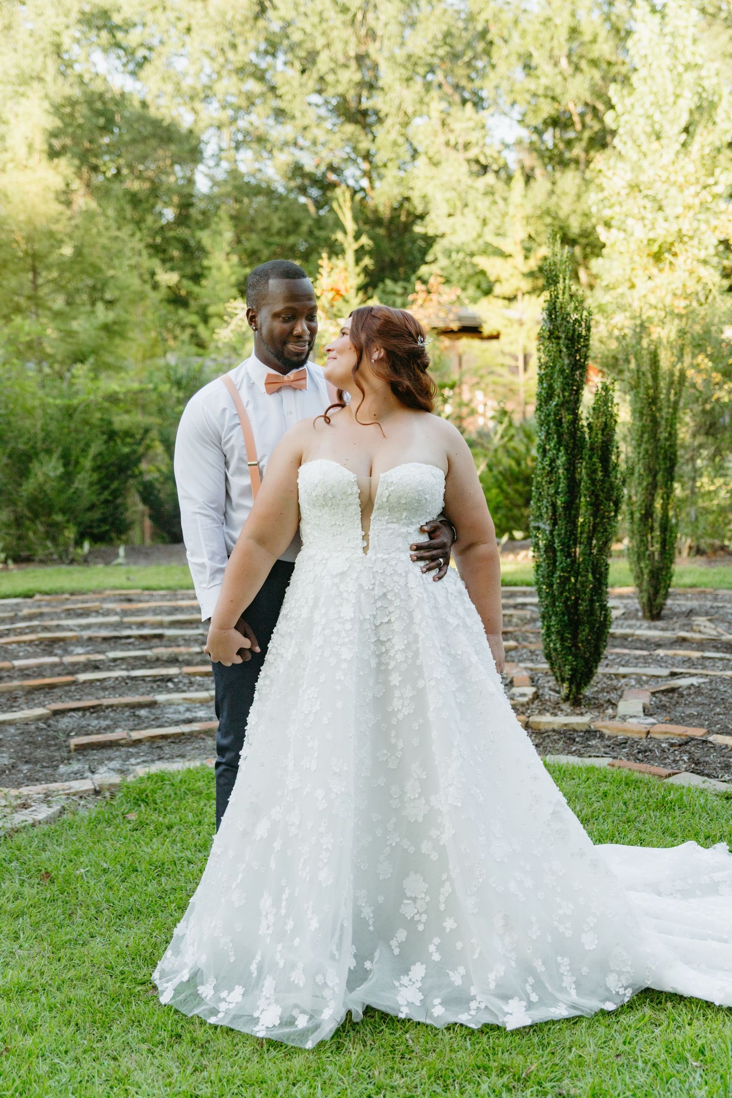 Bride in floral lace ball gown and groom in gray suit with pink bow tie pose in garden amphitheater.