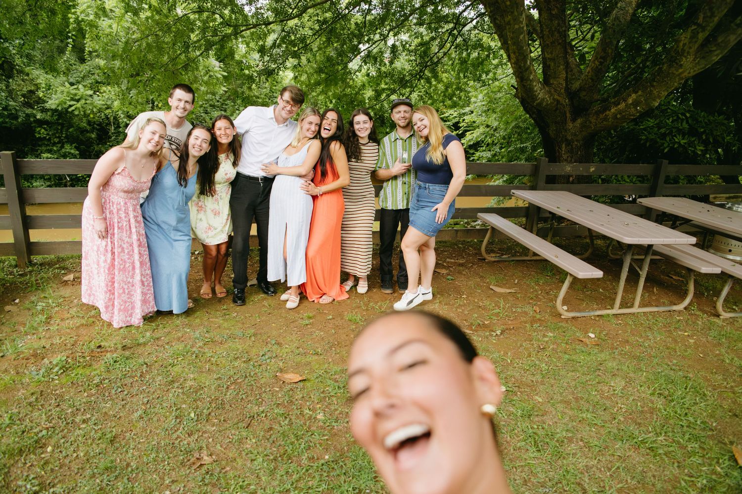 Group of friends smile together outdoors at park gathering, with woman taking smiling selfie in foreground.