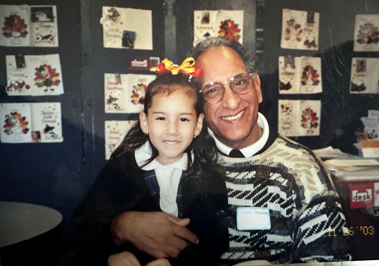 Young girl with yellow bow poses with smiling older man in patterned sweater in front of colorful holiday cards display.
