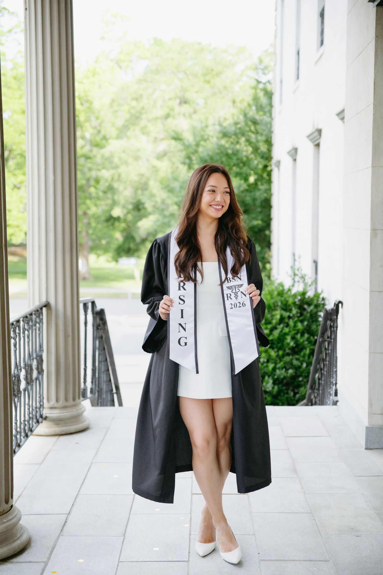 Nursing graduate smiling in black gown and white dress with graduation stole outdoors.
