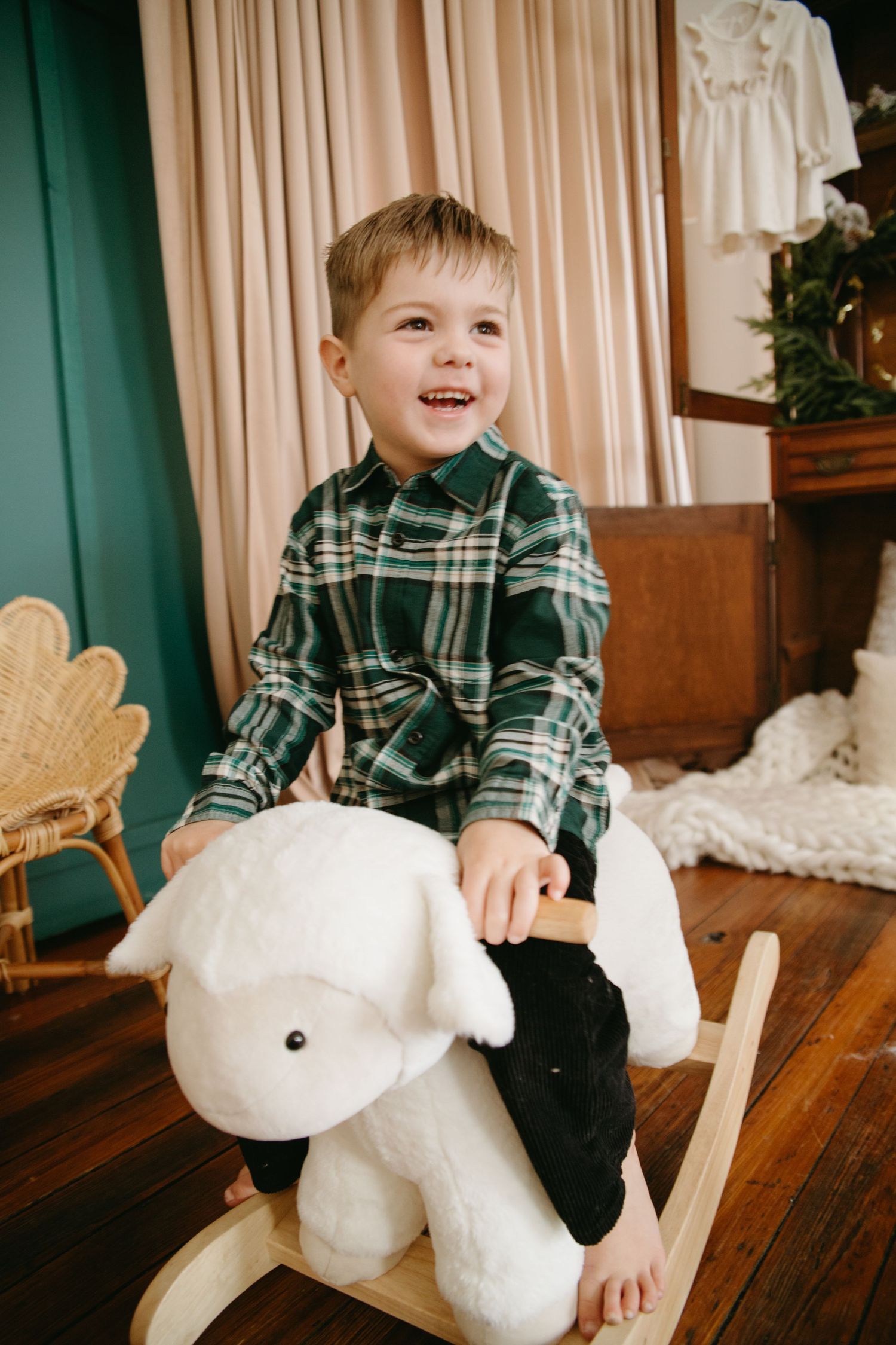 A laughing young boy in a green plaid shirt sits on a white lamb rocking toy in a warmly decorated holiday room.
