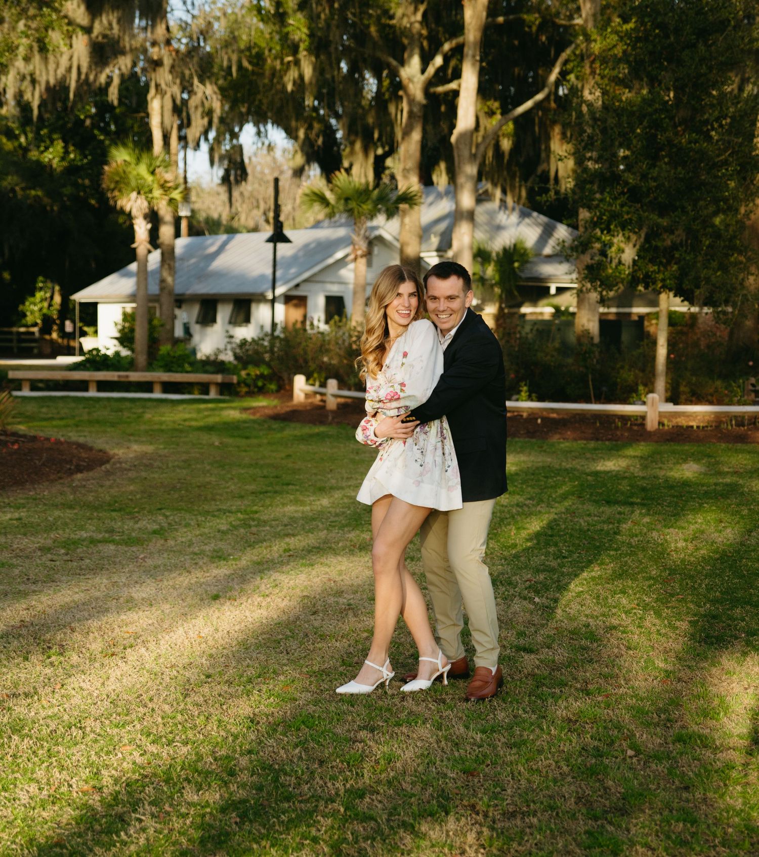 Happy couple embracing and smiling outdoors under Spanish moss trees near a white cottage.