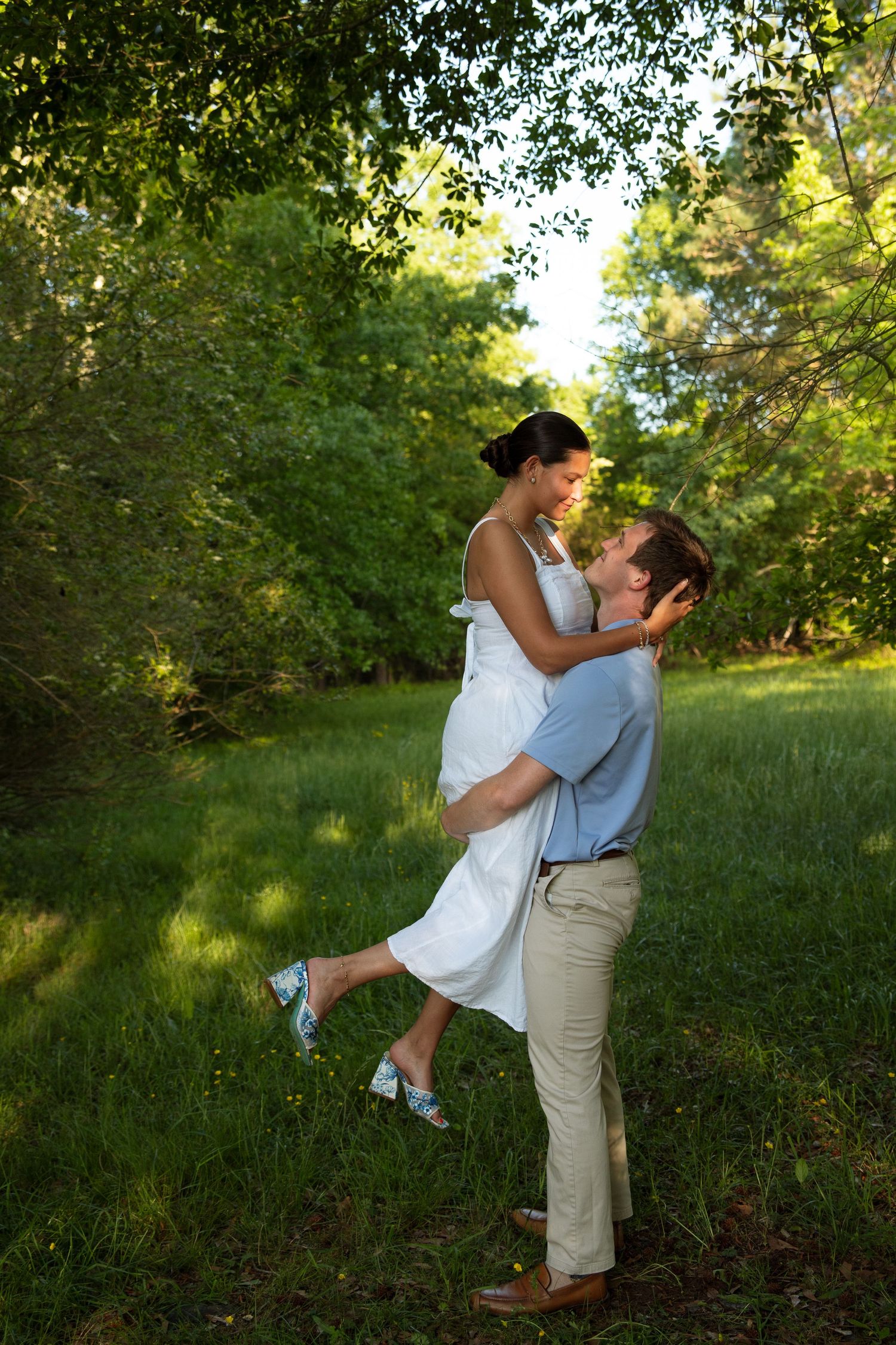 Man lifting woman in white dress in a lush green meadow surrounded by trees in warm evening light.
