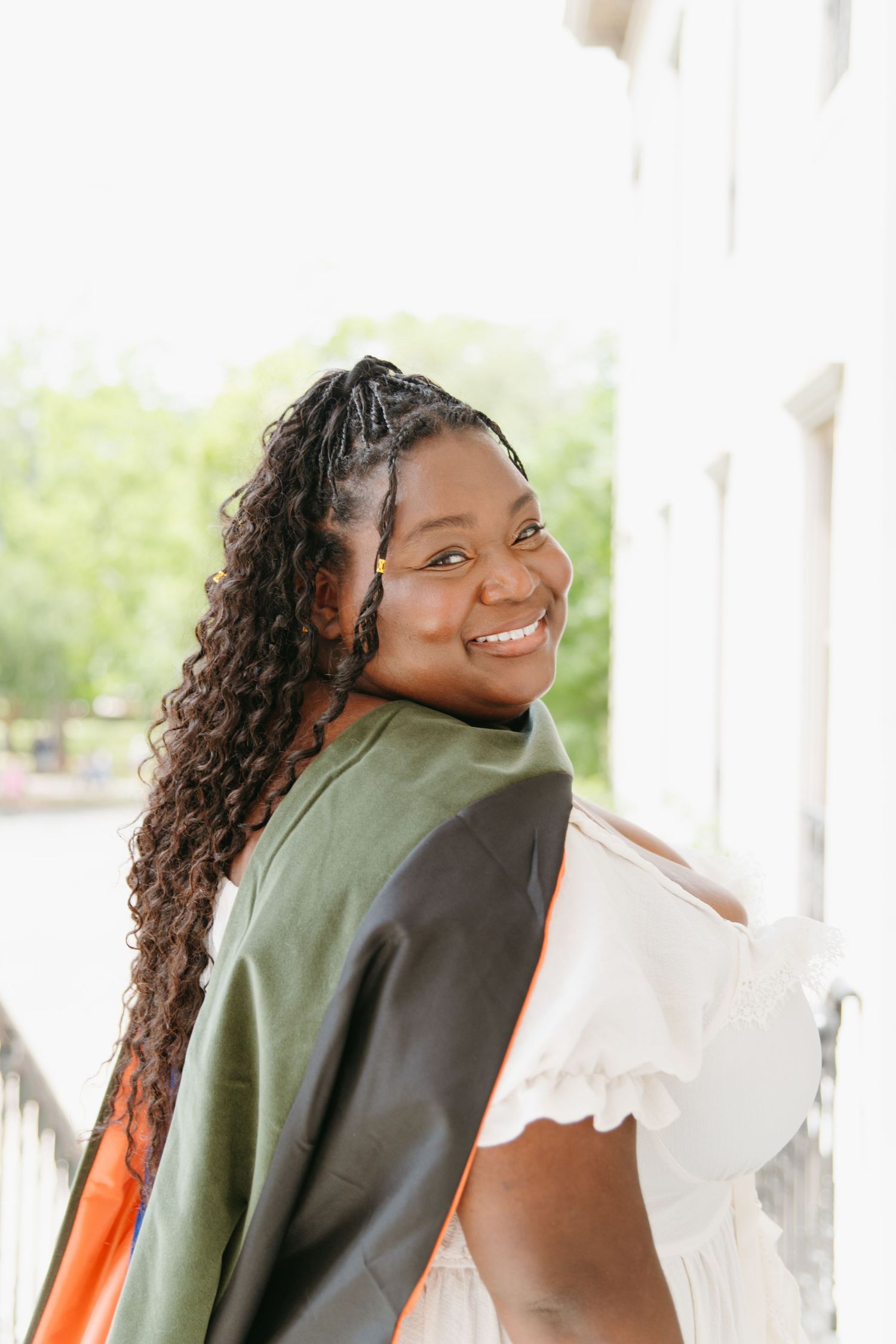Smiling Black woman in white dress with green graduation stole posing outdoors.