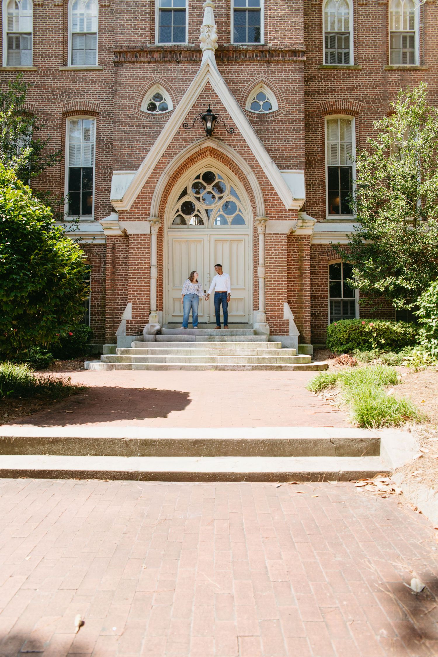 Couple standing together at the entrance of a historic Gothic brick building on a sunny day.