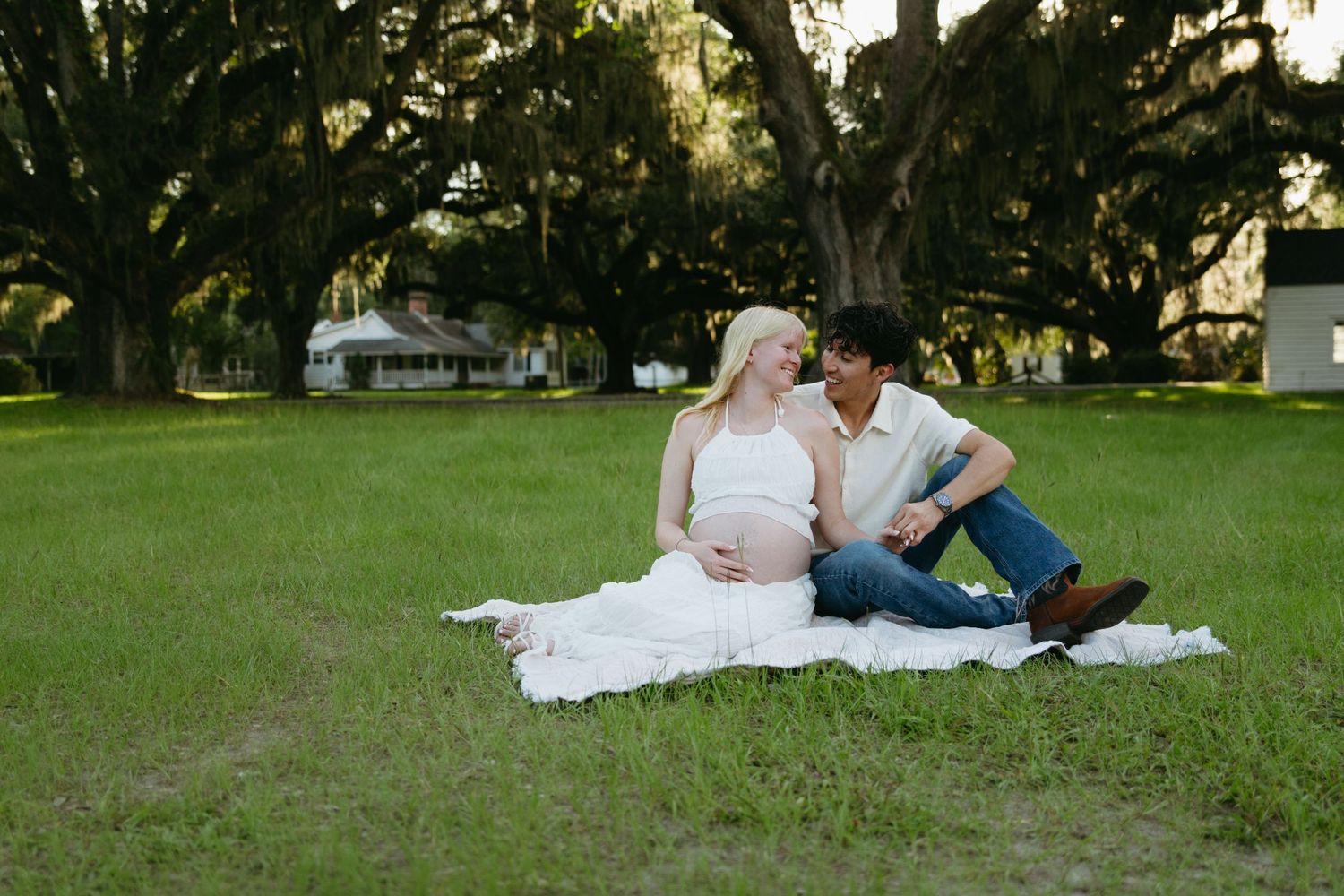 Expectant couple sharing a tender moment on a blanket in a scenic green park setting.