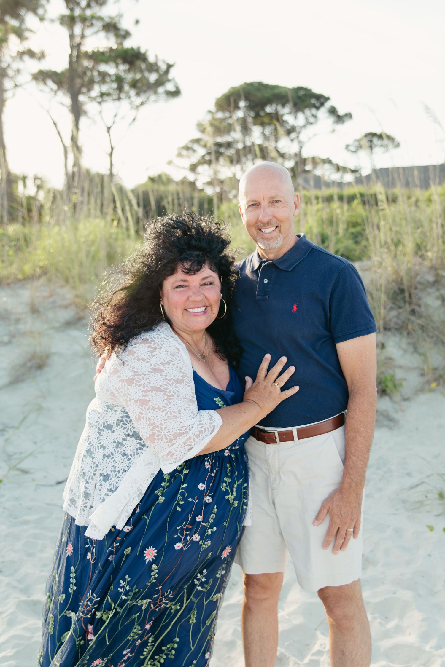 Smiling mature couple embracing on sandy beach dunes with sea grass and pine trees behind them.