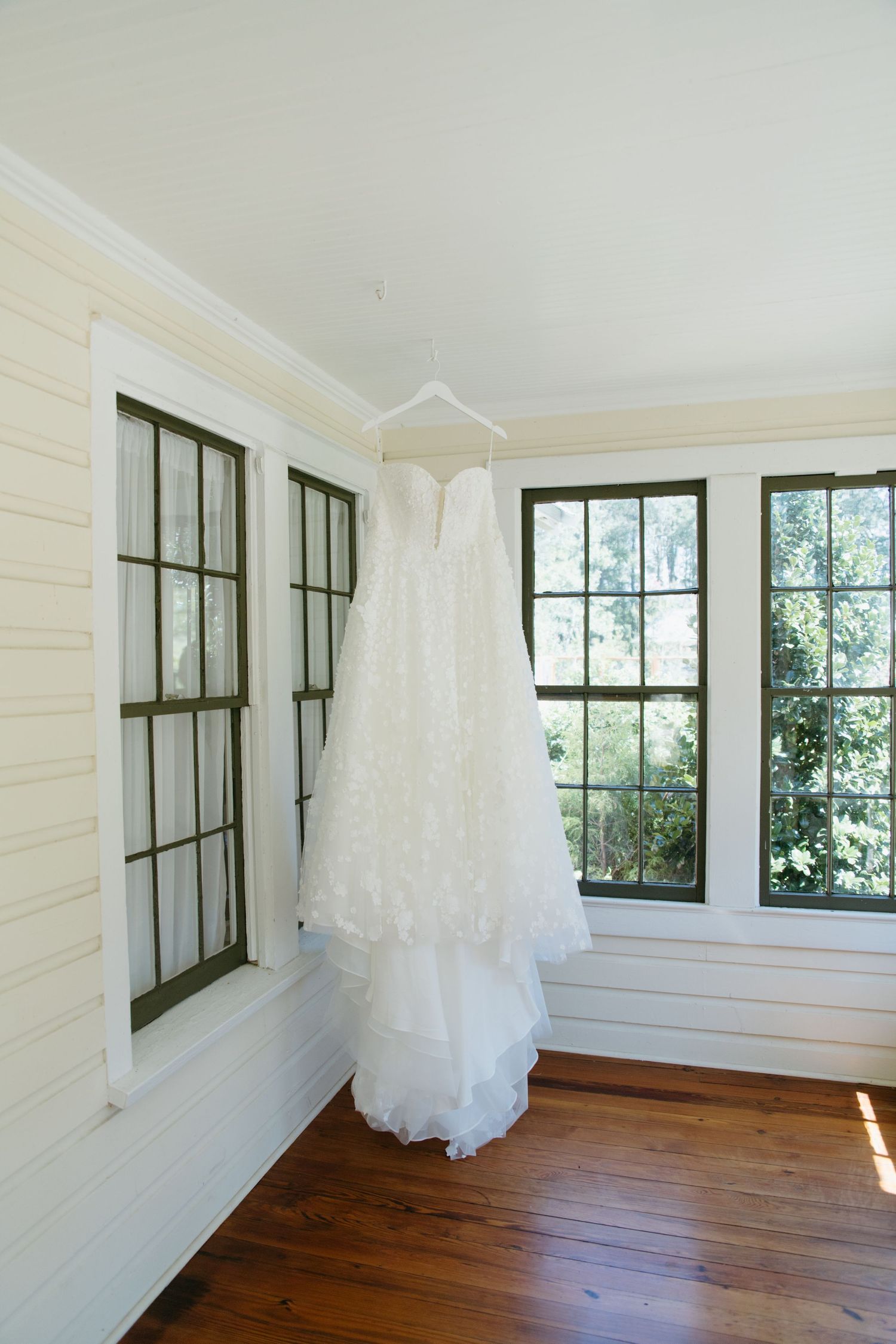 Elegant strapless lace wedding dress hanging on hanger in sunlit room with black-framed windows.