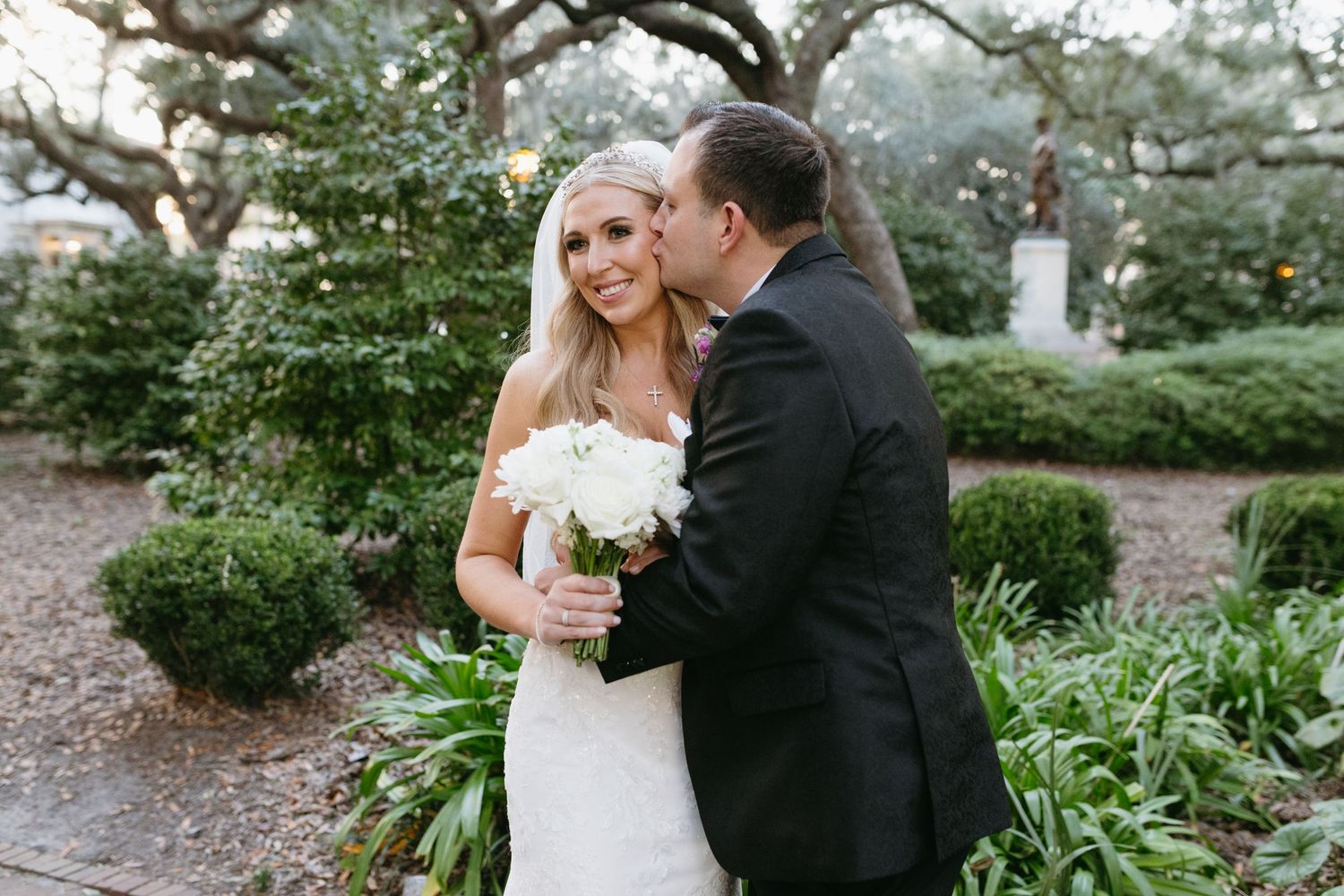 Groom kissing bride on cheek as she smiles holding white bouquet in garden setting.