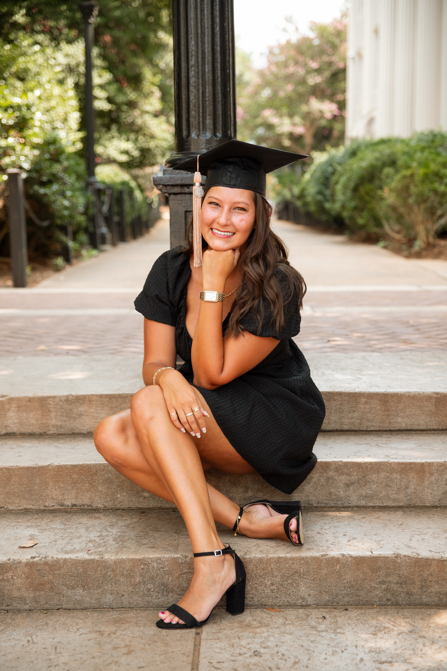 Smiling female graduate in black dress and cap sitting on stone steps for graduation portrait.