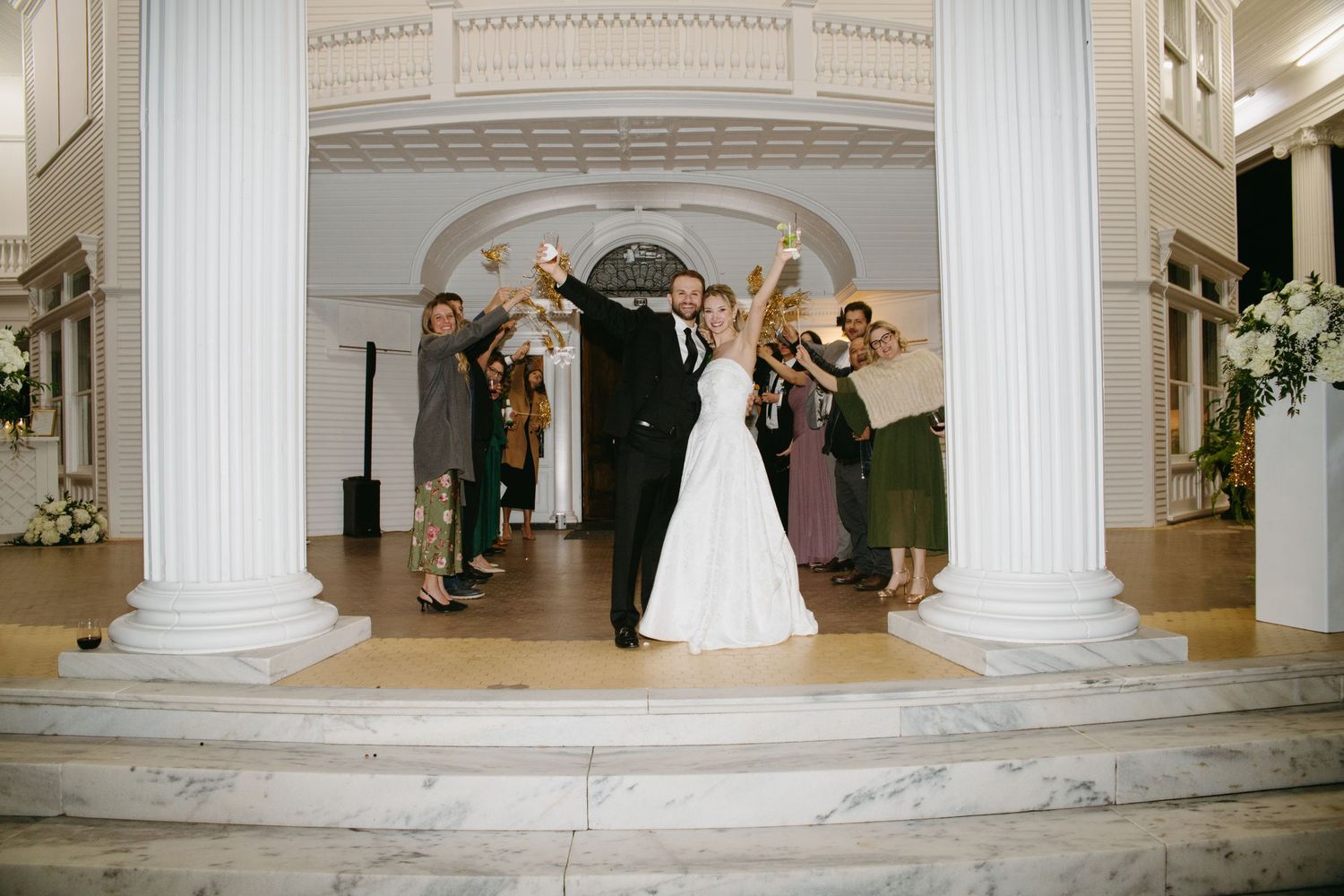 Bride and groom celebrate on steps of elegant venue with white columns as guests cheer around them.