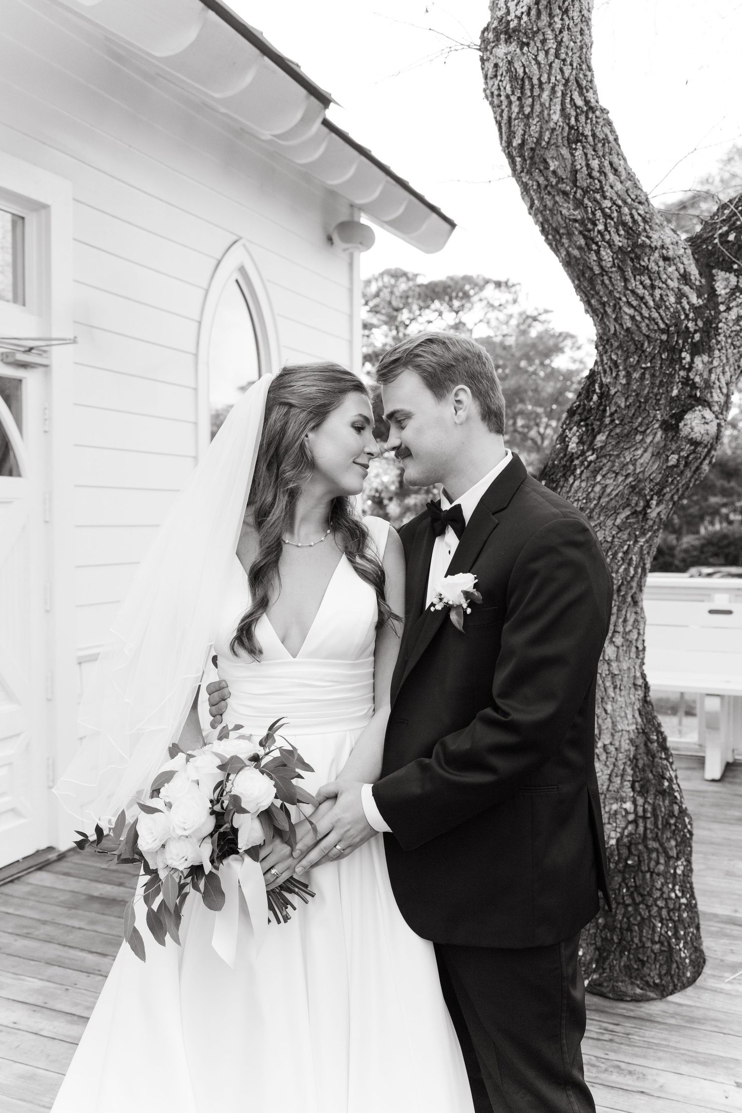 Black and white portrait of bride and groom touching foreheads outside white chapel.