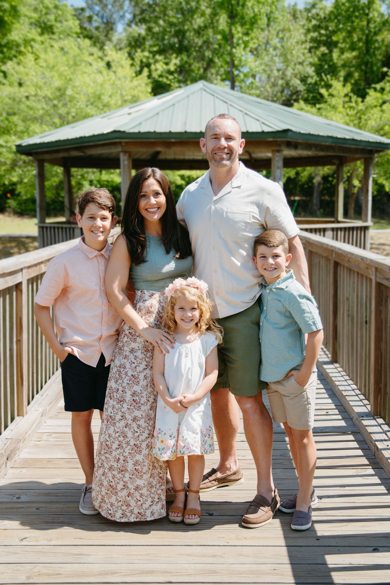 Family of five poses on a wooden bridge near a gazebo in a sunny park.