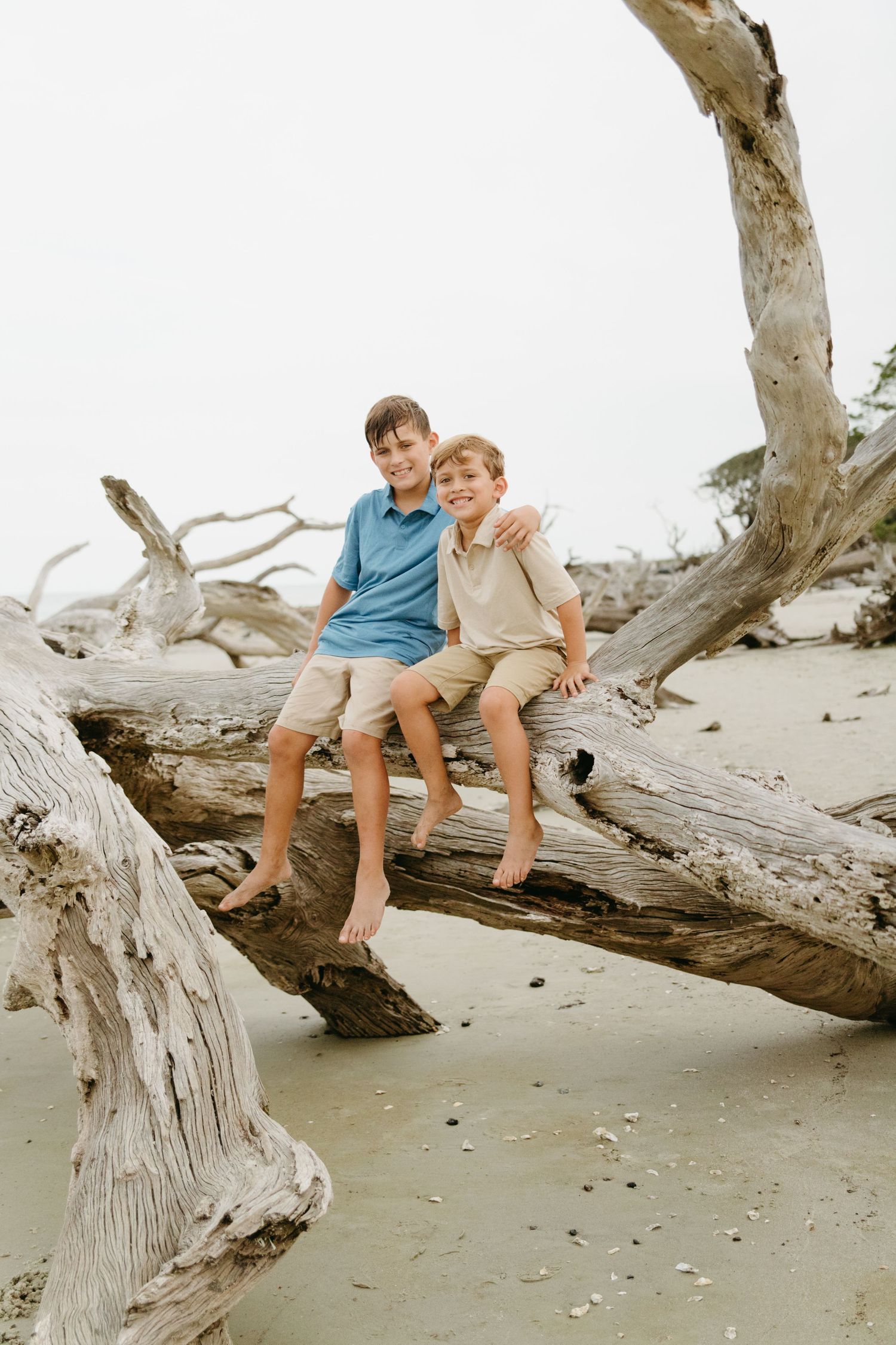 Two boys sit smiling on driftwood at a beach, one with arm around the other.