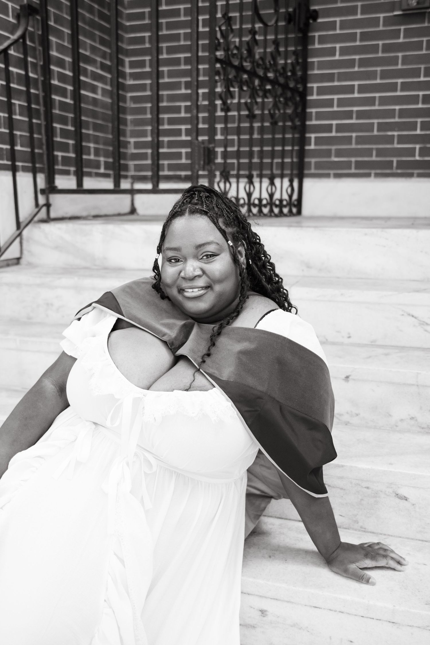 Black and white photo of smiling woman in white dress with graduation stole on steps.